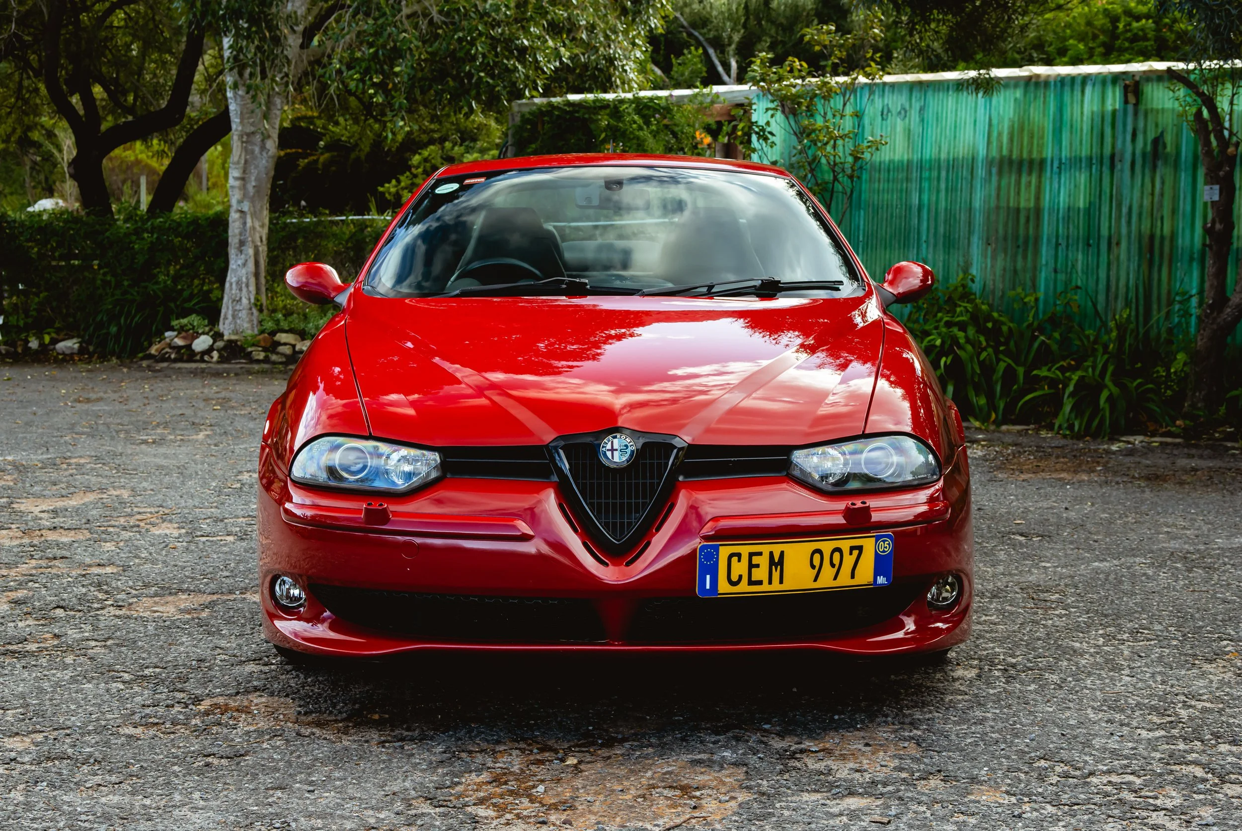 Front view of a red Alfa Romeo car parked on a gravel surface with trees and a green fence in the background.