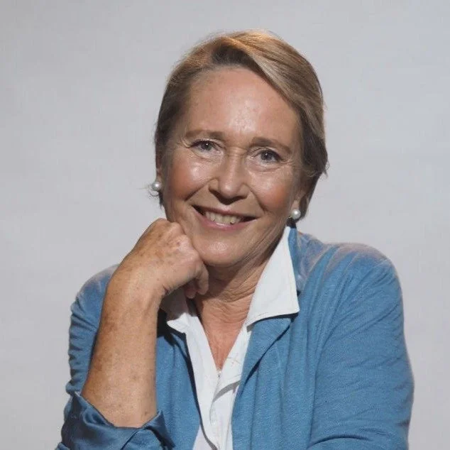 Ritratto di una donna anziana con capelli castani, indossa una giacca blu e una camicia bianca, sorride e si appoggia al suo mento con la mano.