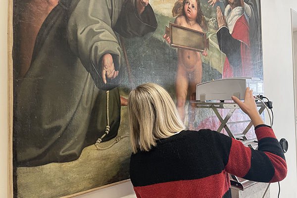 Una persona con capelli biondi chiari che indossa un maglione con lati rossi e nero osserva un dipinto storico, sporgendo il volto e le orecchie dall'angolo inferiore sinistro dell'immagine.