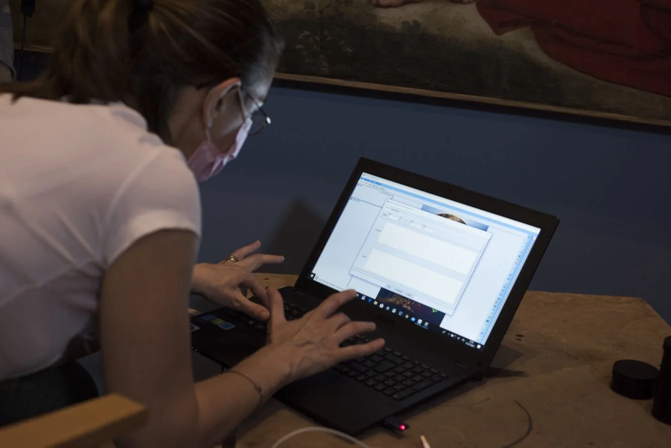 Una donna con capelli raccolti, mascherina, occhiali e braccialetti usa un laptop su un tavolo di legno, concentrata a digitare.
