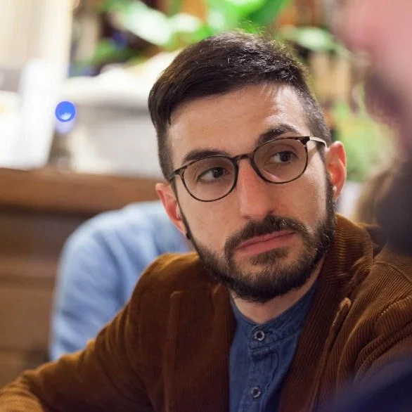 Uomo con barba e occhiali in un ambiente interno, probabilmente un ristorante o un bar, con sfondo di piante e decorazioni in legno.