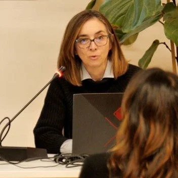 Una donna con occhiali seduta di fronte mentre presenta o parla, con una foglia gigante sullo sfondo.