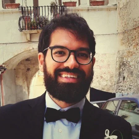 Un uomo sorridente con barba e occhiali indossa un abito con cravatta a farfalla, in un cortile con edifici in pietra e piante in vaso.