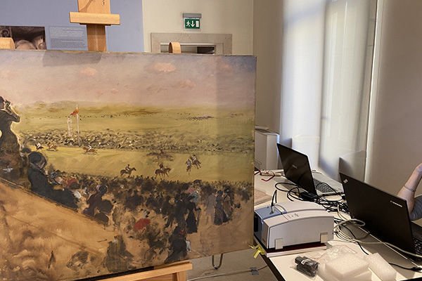 Una grande tela con un dipinto che rappresenta una battaglia storica, collocata su un cavalletto in una stanza con scrivanie e computer.