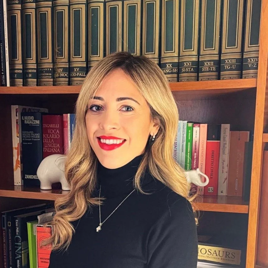 Una donna con capelli biondi, vestita con un maglione nero, che sorride davanti a una libreria con libri di storia, linguistica e altri generi.