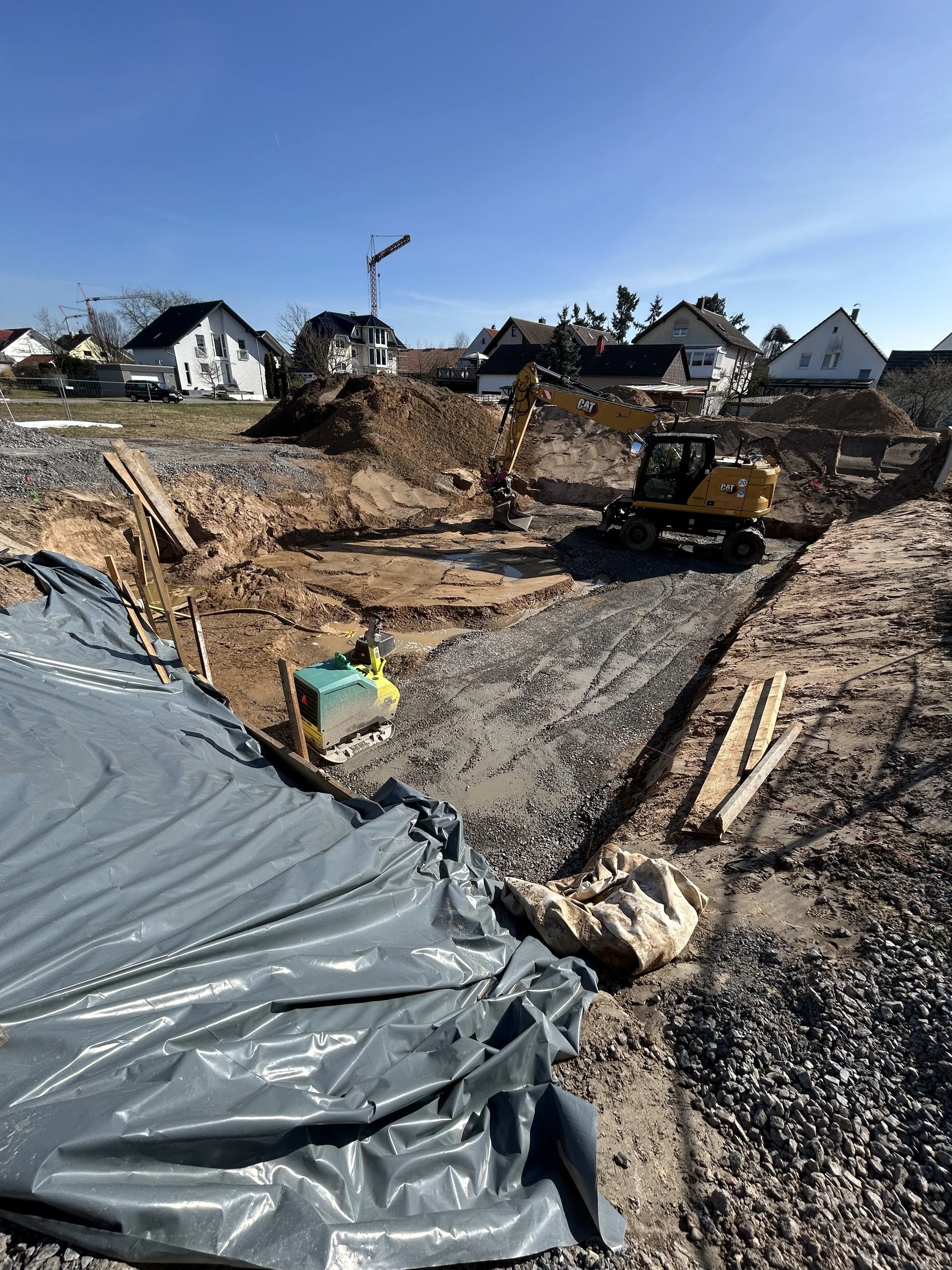 Baustelle mit Bagger, Sandhaufen, Baufolie und Baumaterialien, vor Wohnhäusern bei sonnigem Himmel.