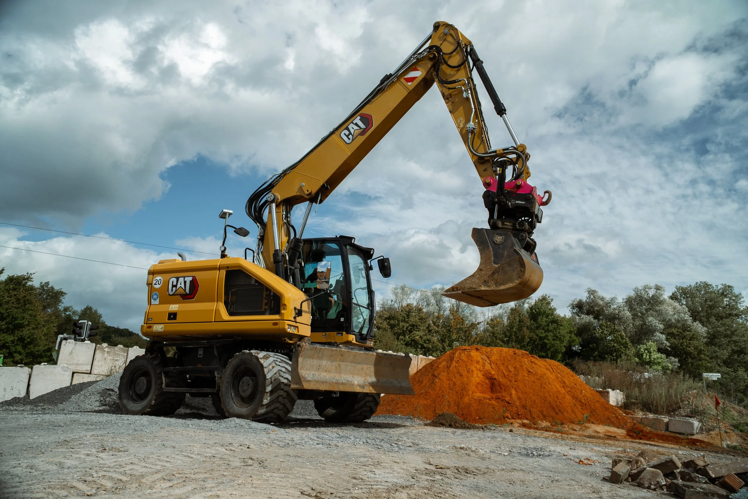 Bagger auf einer Baustelle, hebt Erde über eine kleine Mulde, mit Wolkenhimmel im Hintergrund.
