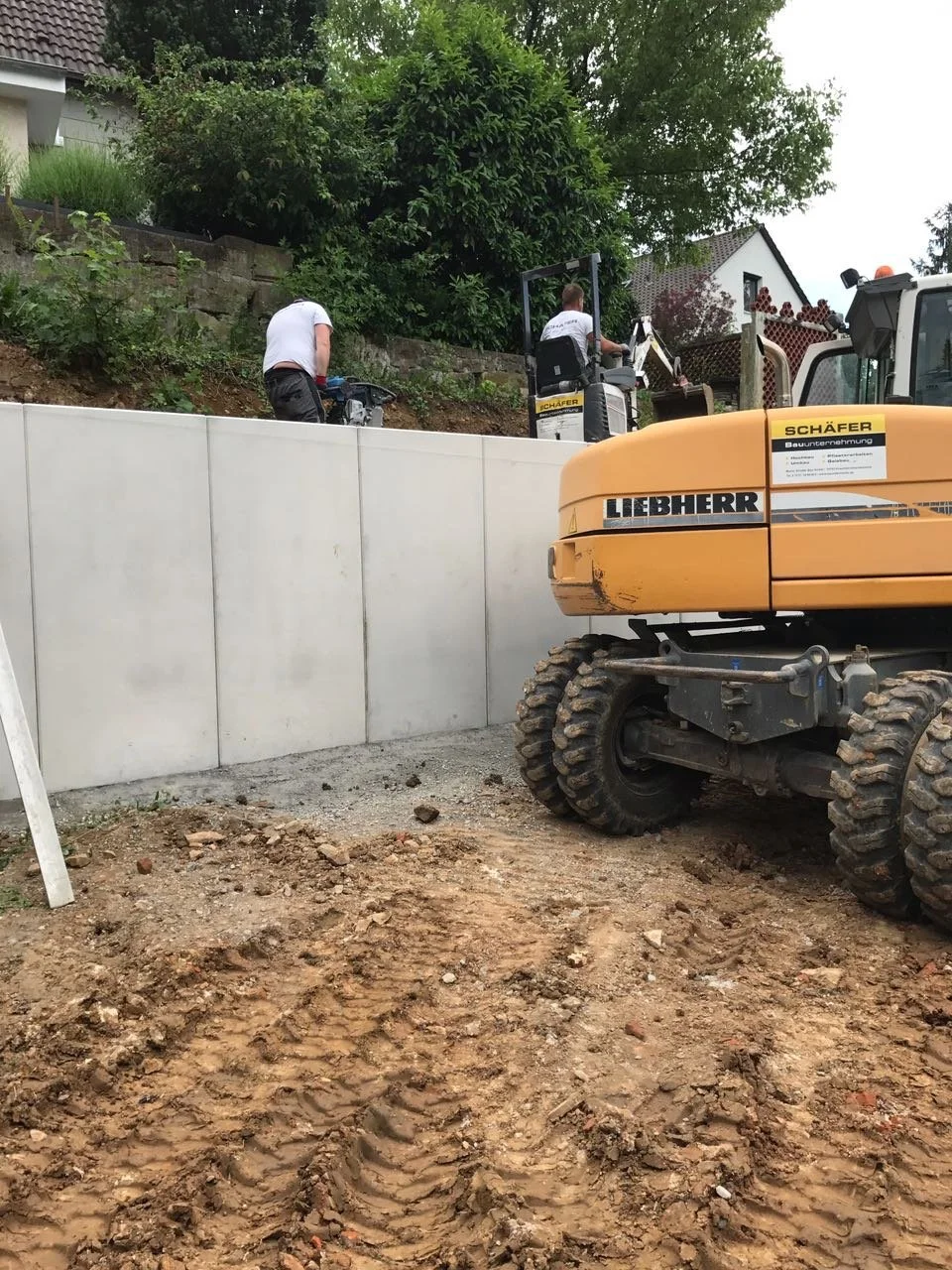 Bauarbeiter bei Bauarbeiten an einer Granitmauer, mit Bagger im Vordergrund auf einem unbefestigten, erdigen Boden.