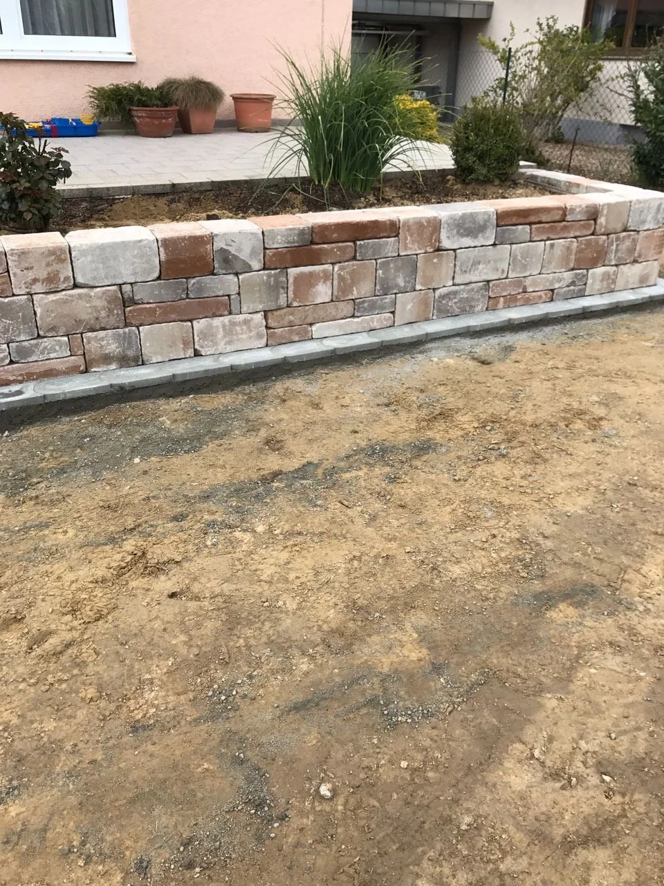 Gartenmauer aus Ziegelsteinen vor einem Haus, im Hintergrund Pflanzkübel mit Pflanzen und eine Terrasse.