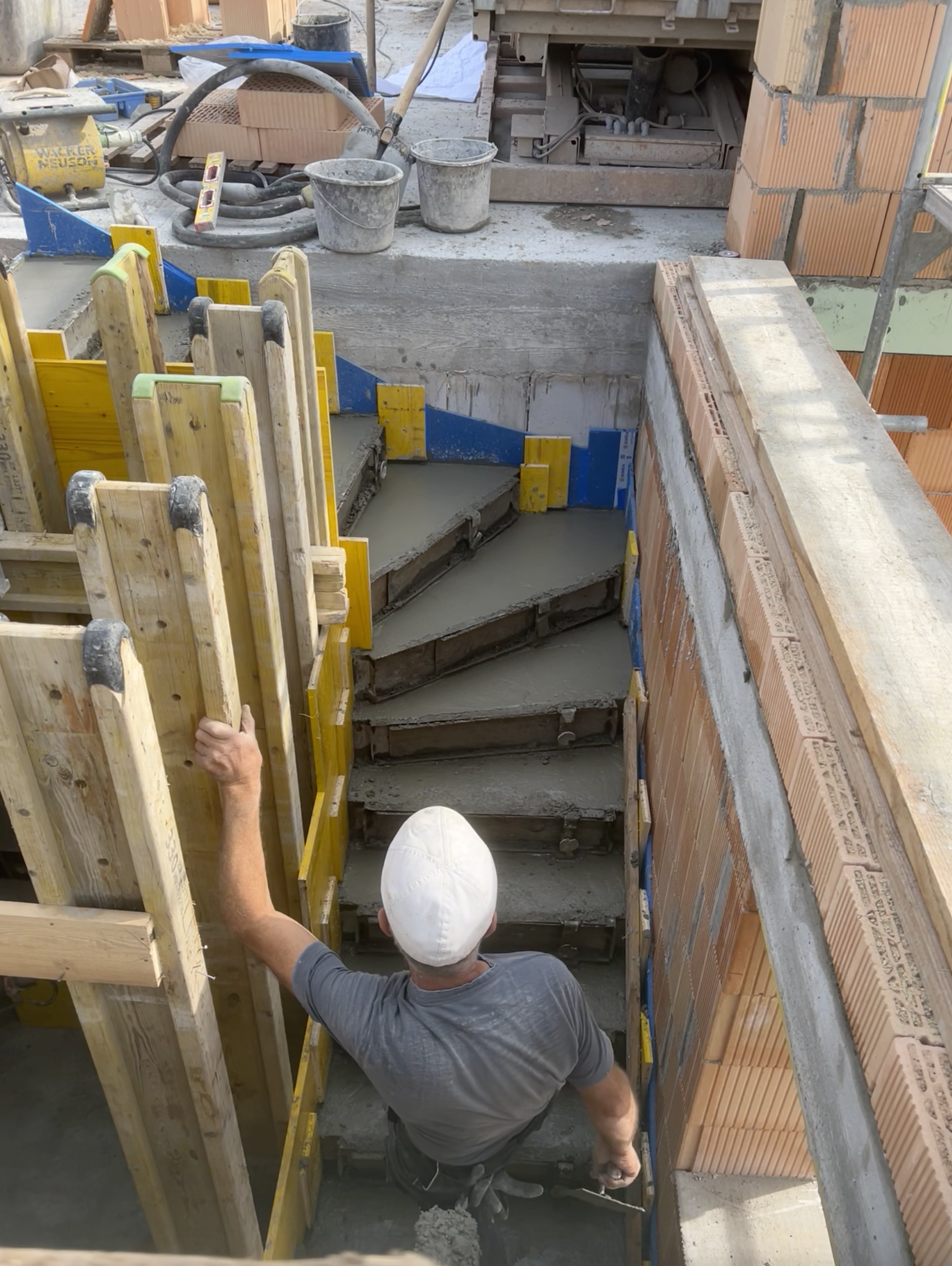 Ein Handwerker baut eine Treppe aus Beton auf einer Baustelle, umgeben von Baugerüsten, Zementsäcken und Baumaterialien.