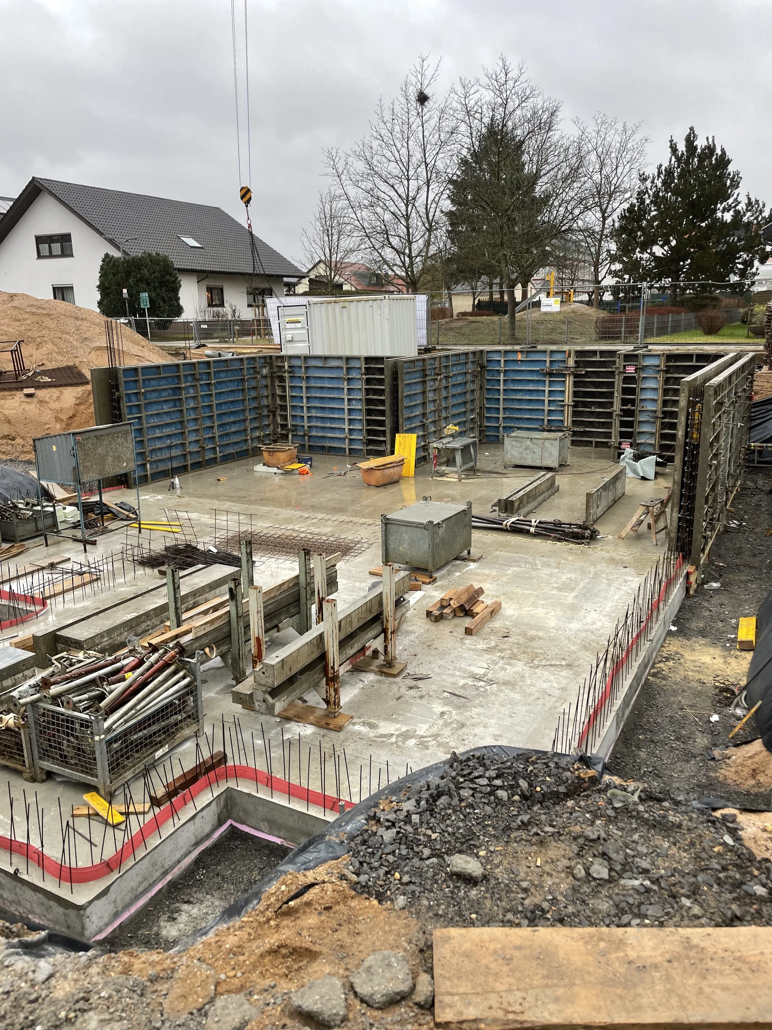 Baustelle mit Fundamenten und Baugerüsten, Wohnhäuser im Hintergrund, bewölkter Himmel.