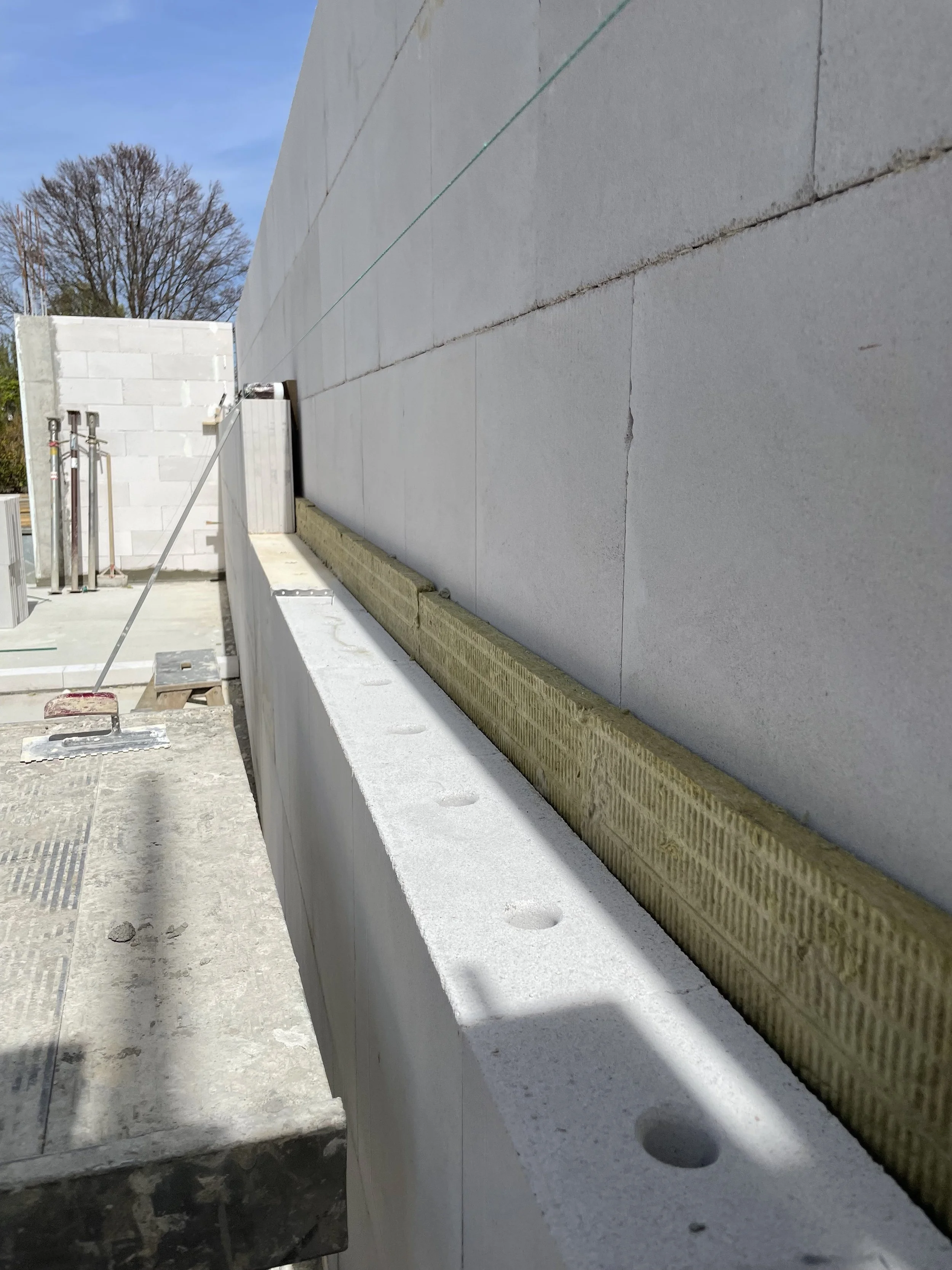 Baustelle mit angemessenen Bauarbeiten, weiße Wand aus Ziegeln, Bauwerk in Entstehung, Werkzeuge und Baumaterialien sichtbar, blauer Himmel.