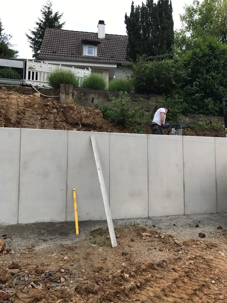 Ein Bauarbeiter arbeitet auf einer Baugrube mit einem Hochwasserschutzwall. Im Hintergrund ist ein Haus mit einer Terrasse und Bäumen zu sehen.