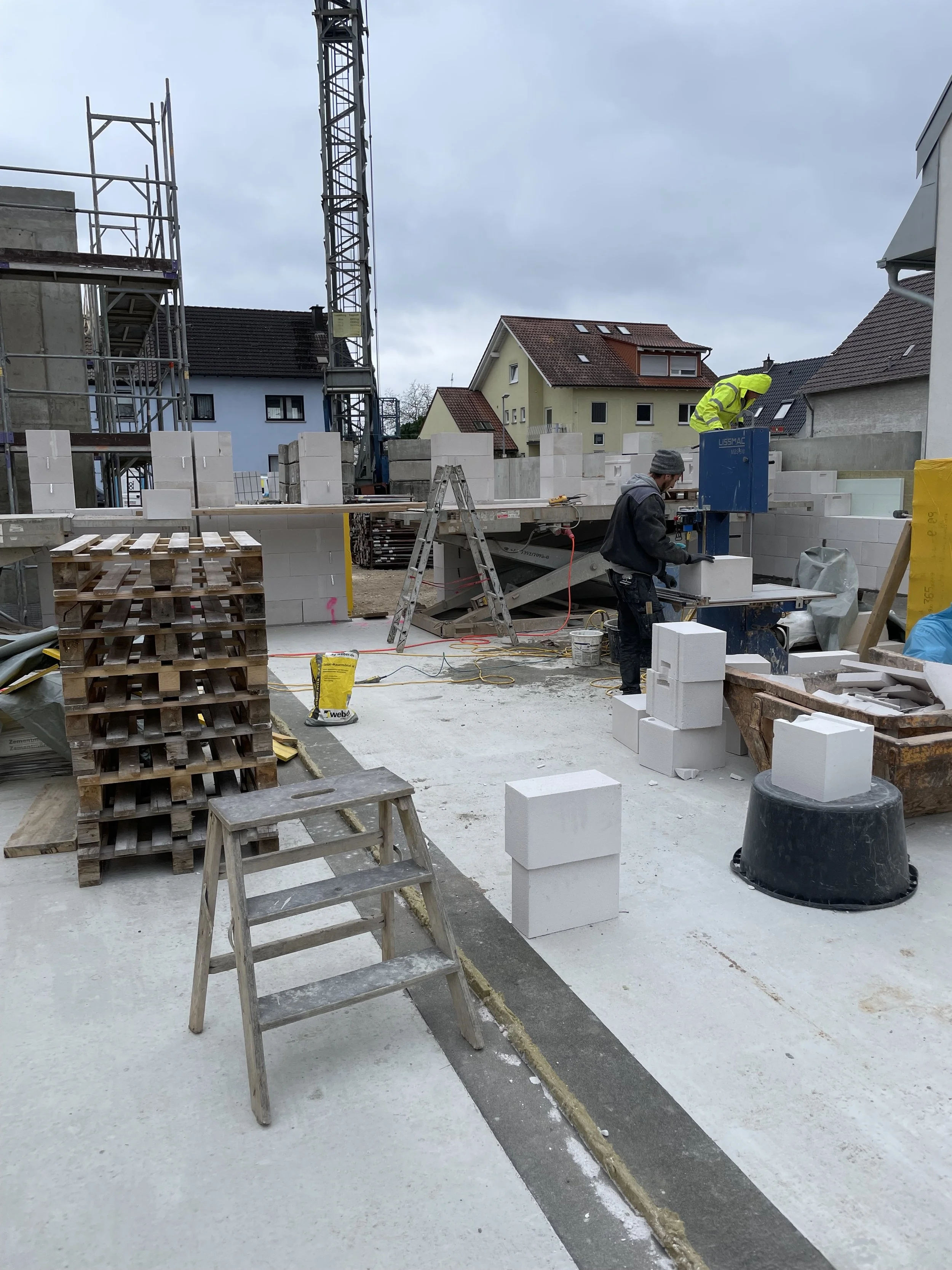 Bauarbeiter arbeiten auf einer Baustelle, dort sind Ziegelsteine, Leitern und Baugeräte sichtbar, im Hintergrund stehen Häuser und ein Kran.