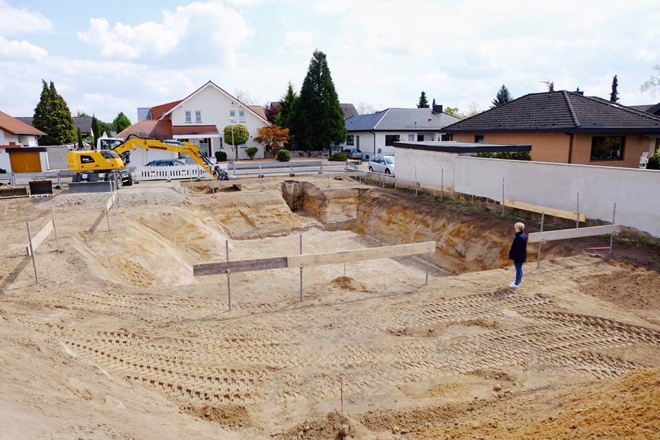 A construction site in a residential area with a large excavation, a person observing, and a yellow excavator in the background.