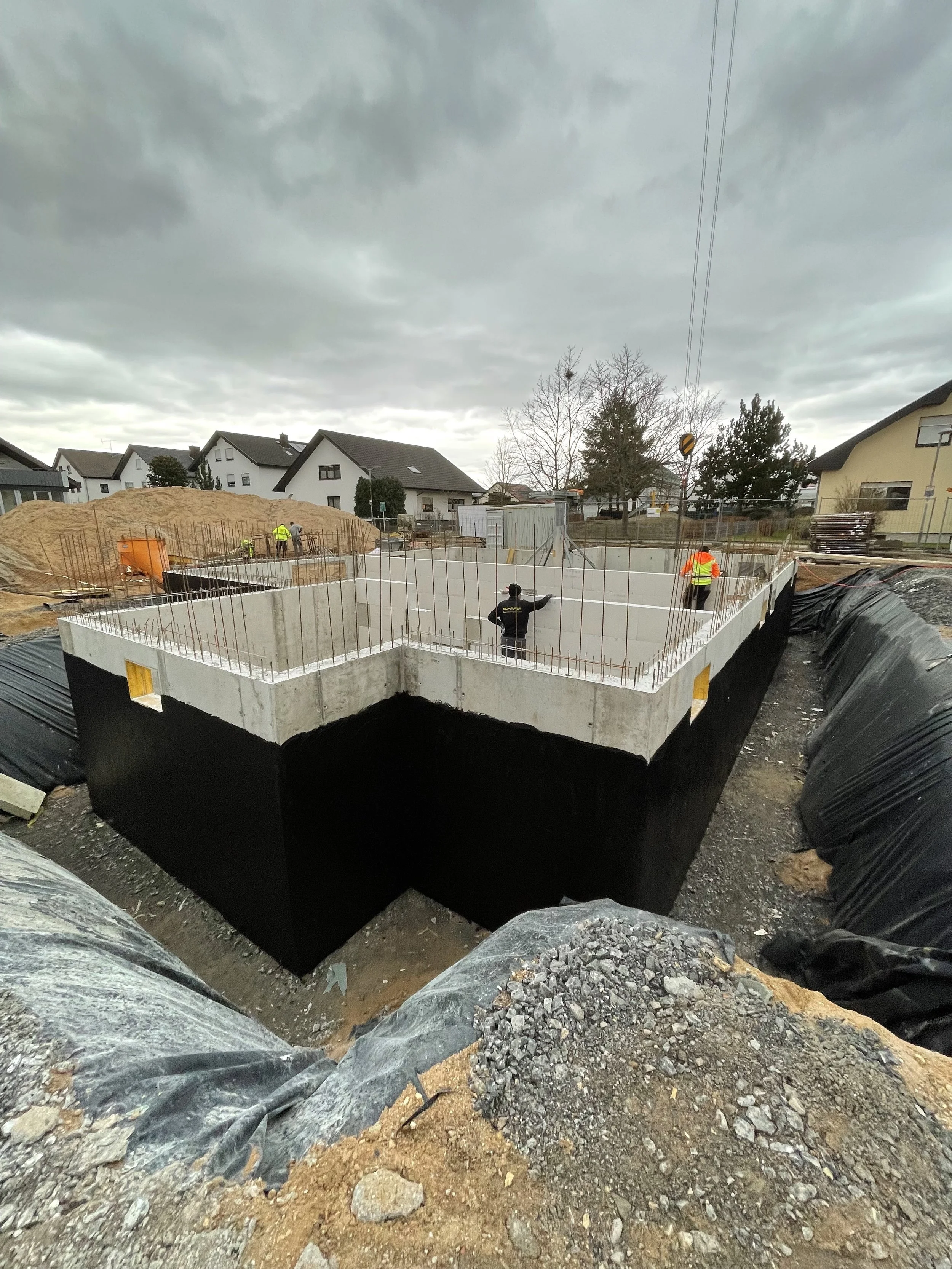 Baustelle mit Betonfundament und Bauarbeitern, im Hintergrund Wohnhäuser, bei bewölktem Himmel.