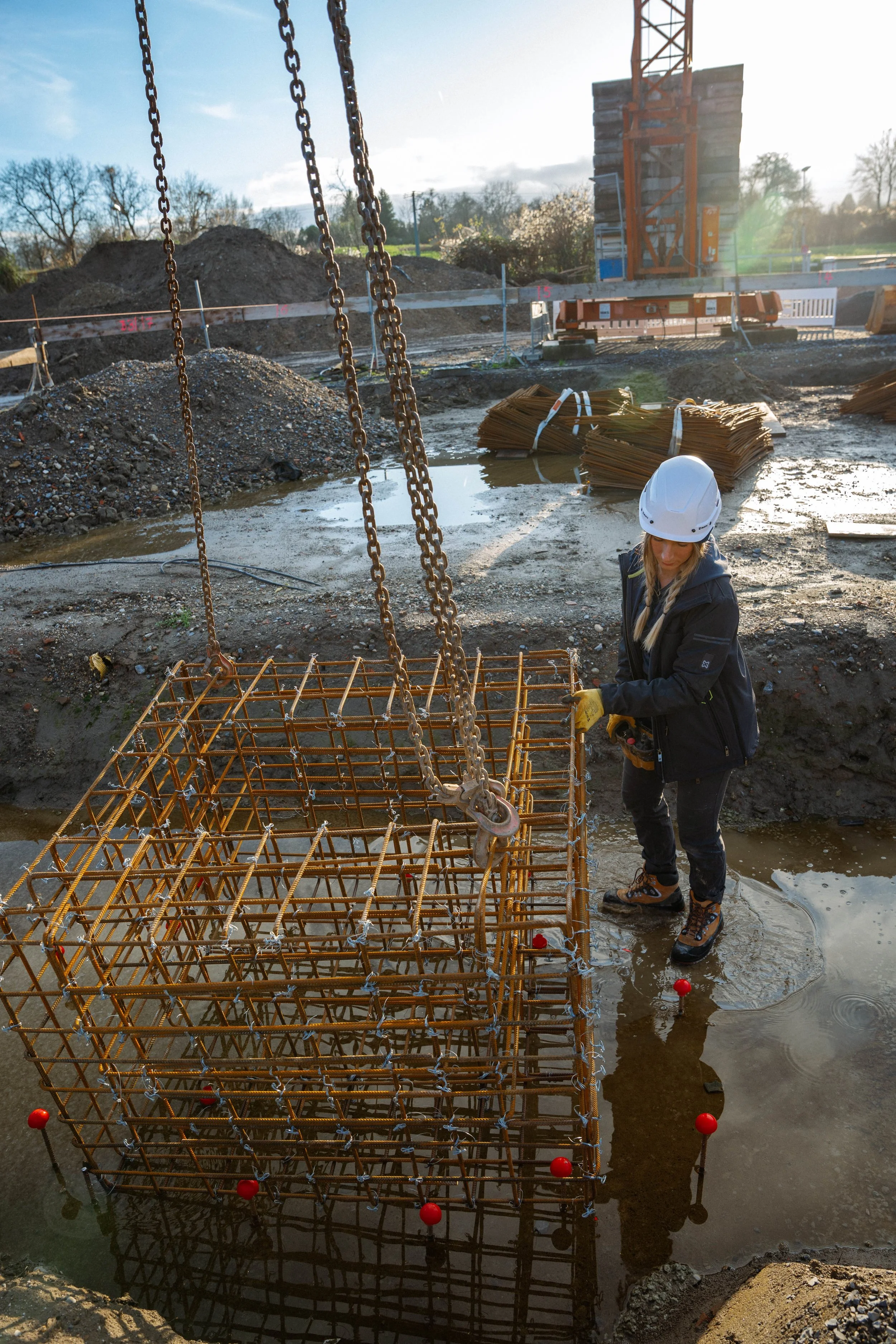 Bauarbeiterin mit Sicherheitshelm arbeitet an Bewehrungskorb auf Baugrube in Außengelände, im Hintergrund Baustelle mit Baugerüst, Erde und Wasser.