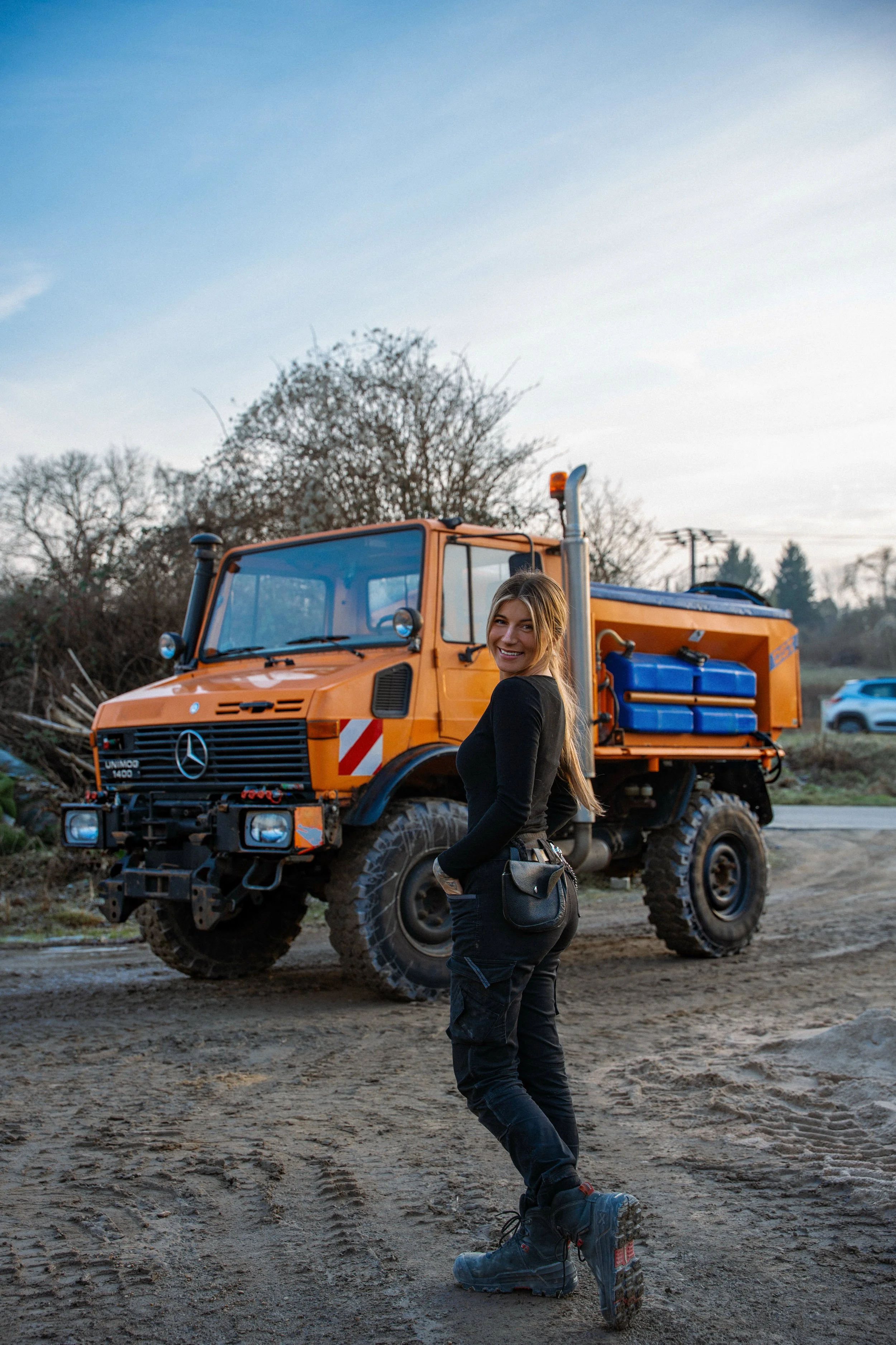 Eine lächelnde Frau steht vor einem orangefarbenen LKW auf einer unbefestigten Straße in einer ländlichen Gegend bei Sonnenuntergang.