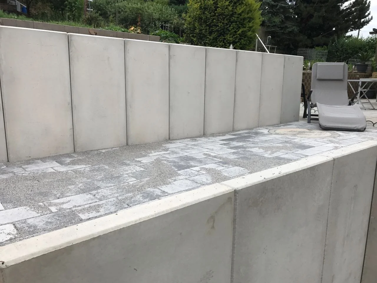 Ein Außenbereich mit weißem Betonmauer, gepflastertem Boden und Liegestuhl im Garten.