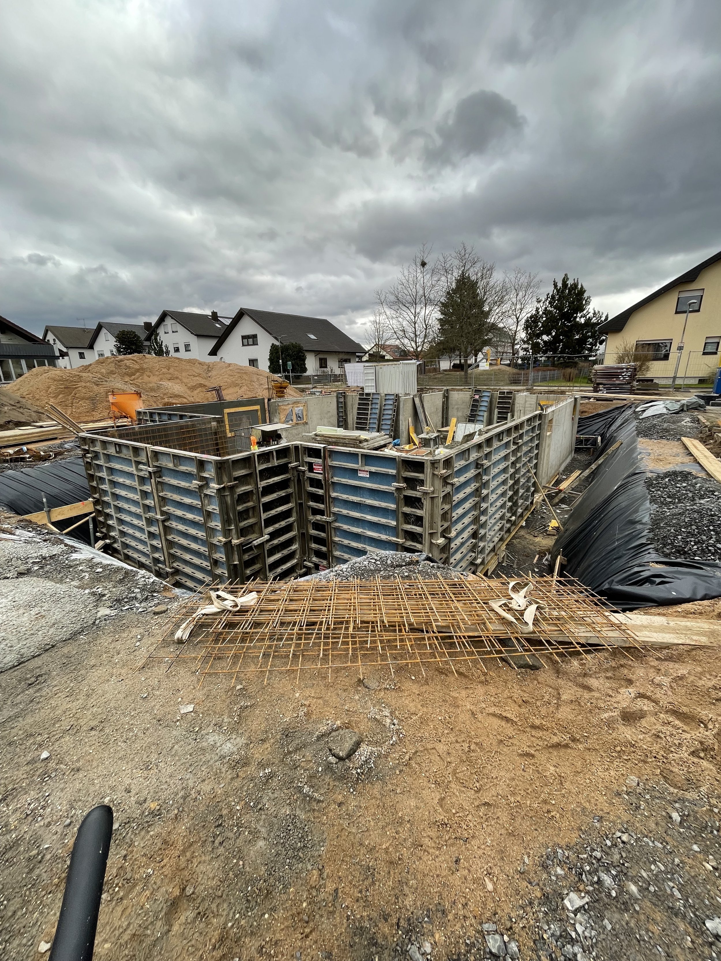 Baustelle mit ausgehobenem Fundament und Baugerüsten, umgeben von Sand und Baumaterialien, bei bewölktem Himmel.