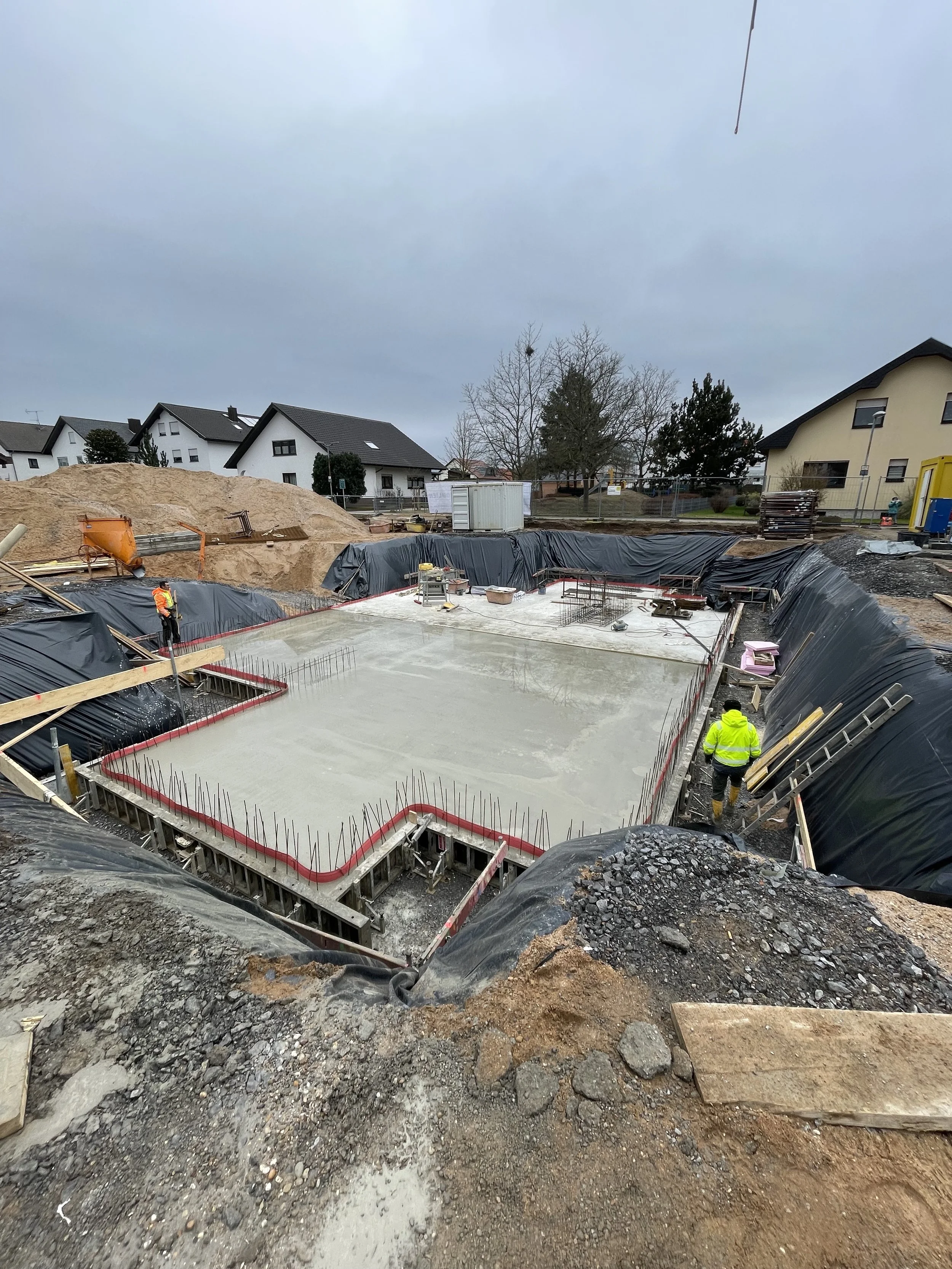 Baustelle mit Betonfundament, Bauarbeiter in gelber Jacke, umliegende Häuser und ein grauer Himmel.