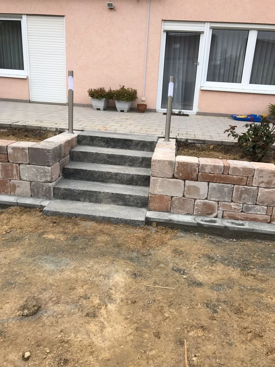 Neue Steinstufen führen zu einer Terrasse vor einem Wohnhaus mit rosa Wand, weißen Fenstern und Tür, sowie Pflanzgefäßen.
