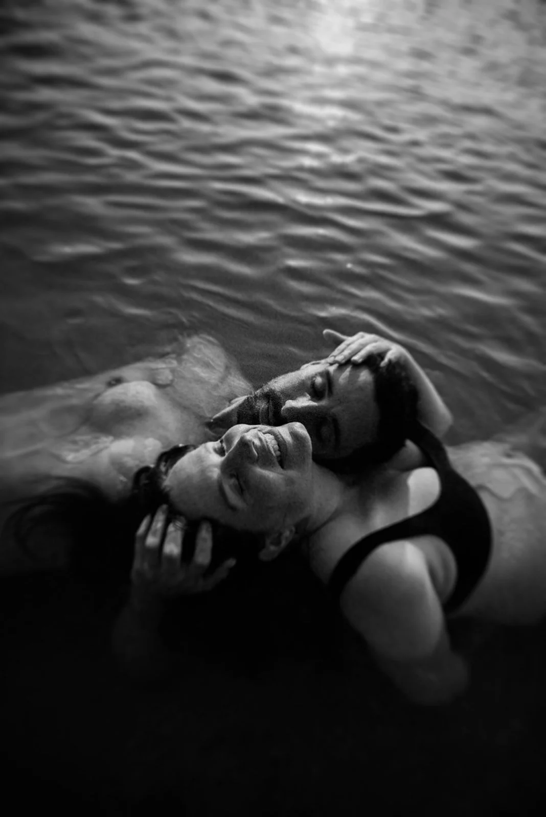 A couple in water, smiling and holding each other closely, with the woman touching the other's face and the man resting his head on his lover's shoulder.