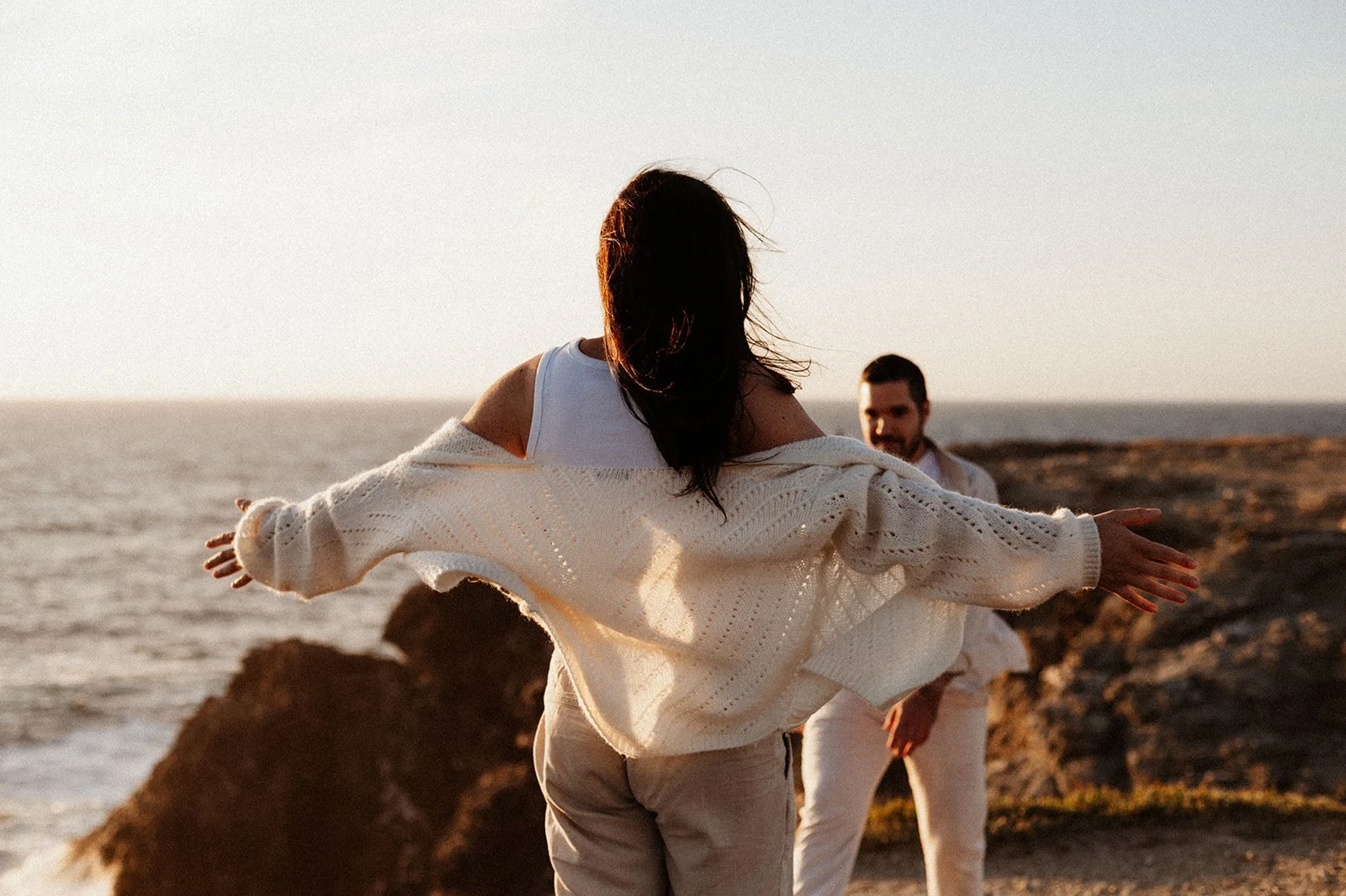 A wide shot of a woman seen from behind, wearing a woollen cardigan, her arms wide open, waiting for her husband to run over and give her a hug, at golden hour on a cliff