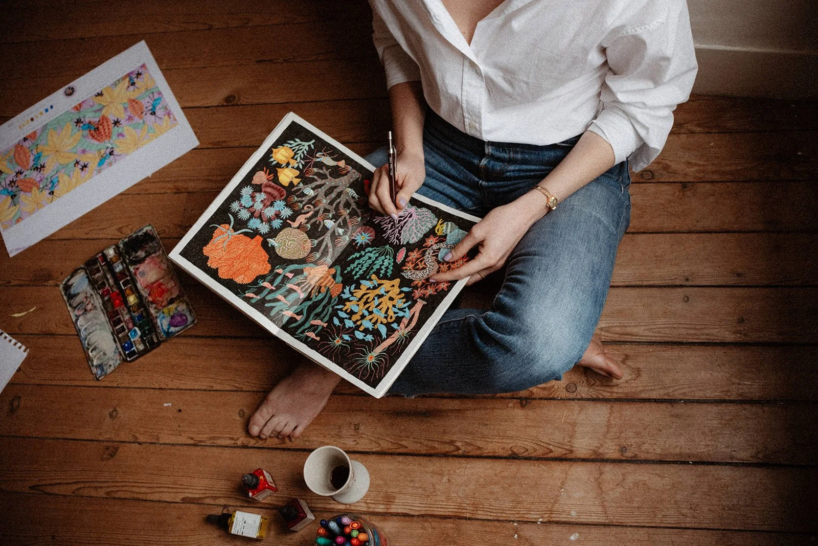 A person sitting cross-legged on a wooden floor, painting a colorful botanical artwork with paints and brushes. There are art supplies, a palette, and artwork on the floor around them.