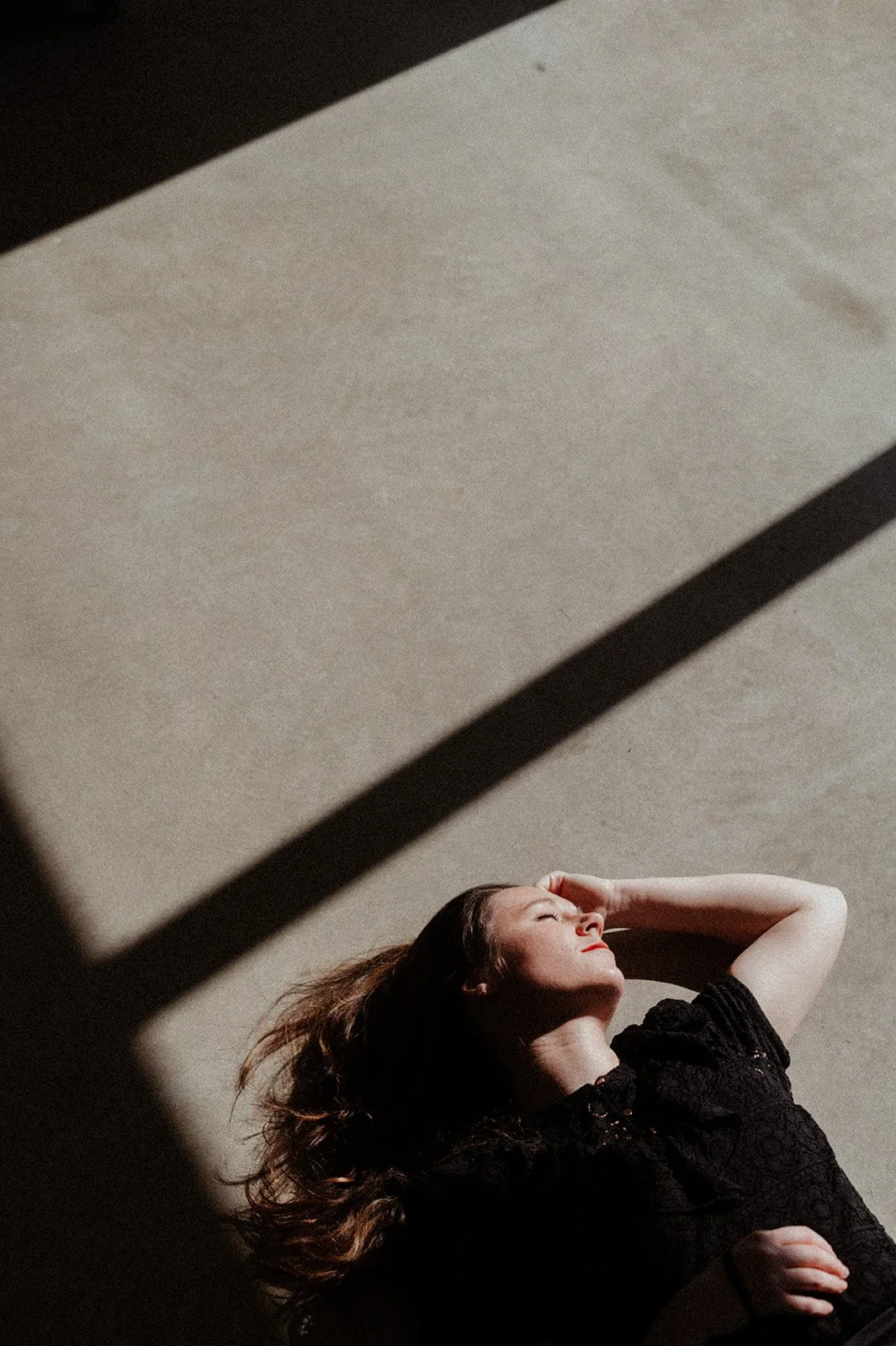 a photo of a young woman lying on a concrete floor, a diagonal rectangle of light cutting across her silhouette, her long brown hair cascading onto the floor, one hand on her stomach and the other resting alongside her face, her eyes closed