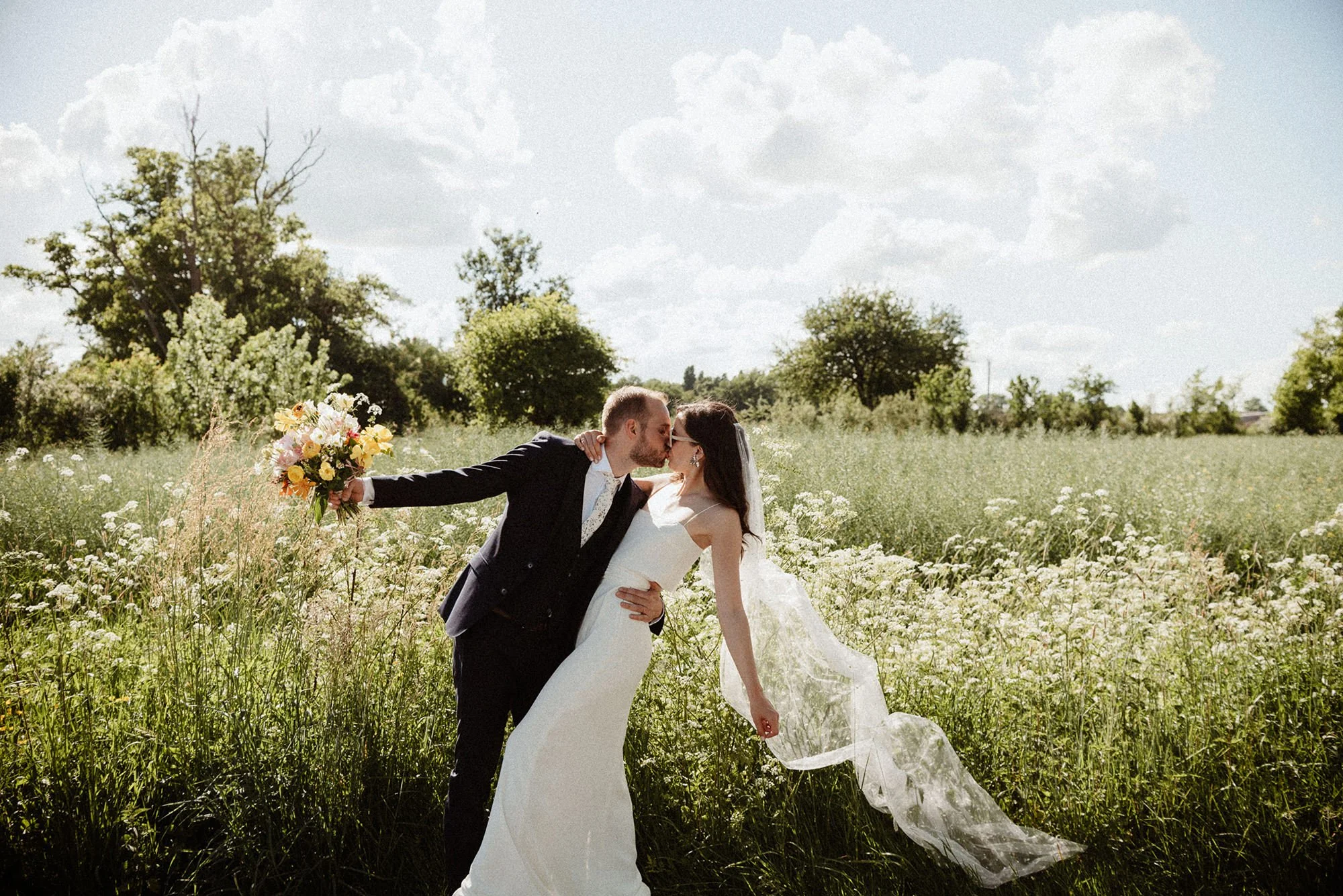 A side view of a newlywed couple: the groom, holding a colourful bouquet with his arm outstretched, kissing his wife, who is leaning back with her veil blowing in the wind, set against a field of flowers, in the style of Wes Anderson