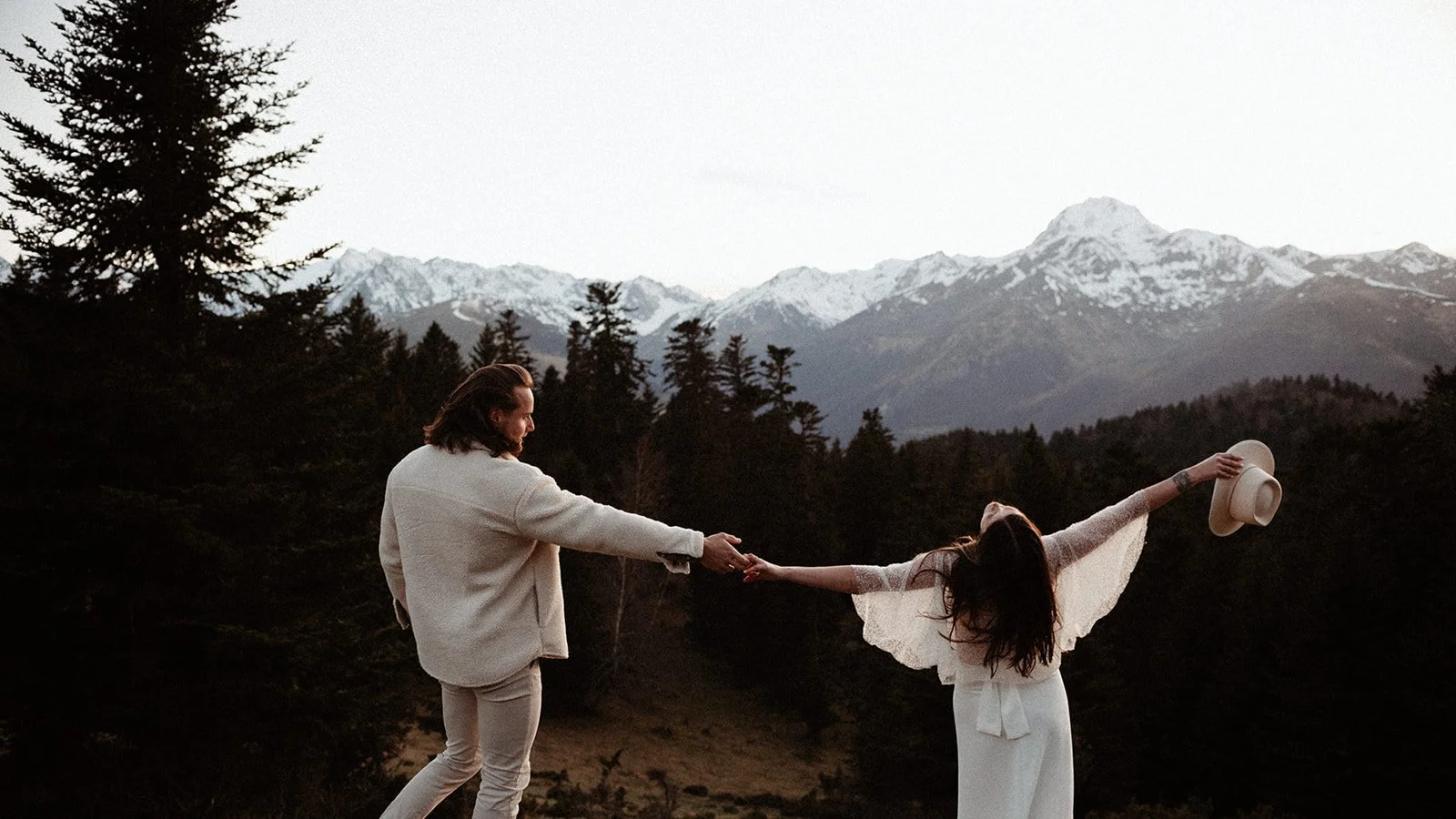 A couple in wedding attire dancing in a wooded mountainous landscape, holding hands, whilst she holds her white hat aloft, with snow-capped peaks in the background, depicted in colour