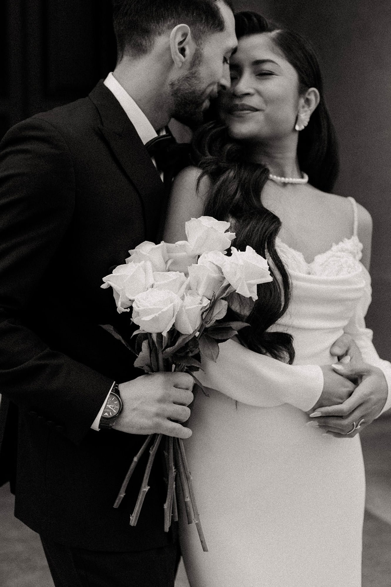 A black-and-white photograph of an elegant, old-money-style bride and groom, with the groom holding a bouquet of white roses and leaning in to kiss his wife, who has long, wavy hair and a pearl necklace, with one hand around his waist