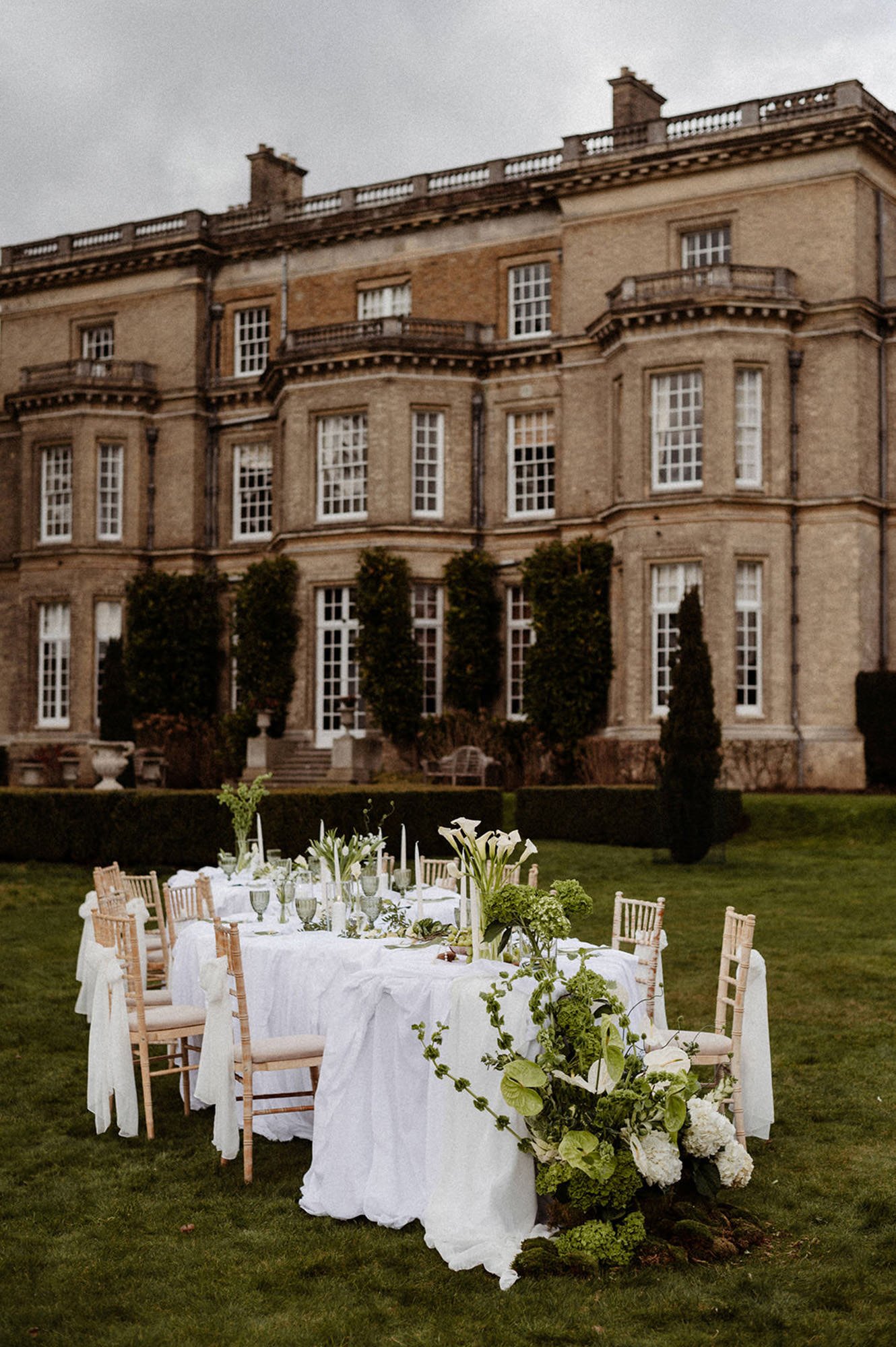 A table set in the grounds of an English manor house in the Cotswolds, with large white tablecloths and white bows on the chairs, adorned with large green and white bouquets, calla lilies, white candles and pearls