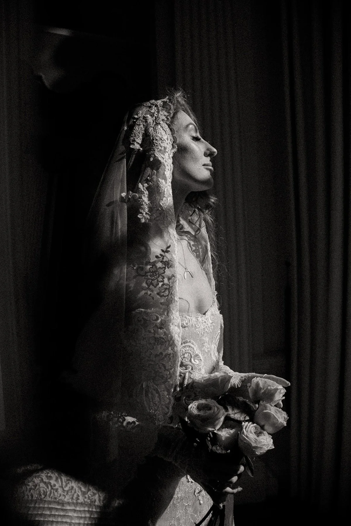 black-and-white photo, taken in profile, of a bride getting ready. She is standing in front of a window, her eyes closed, holding her bouquet; the light illuminates her long, wavy auburn hair and her lace veil; she looks like a Madonna