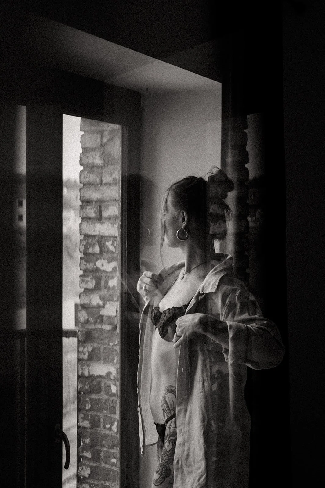 A woman with her hair in a bun, wearing large hoop earrings, standing by a door with a glass window, looking outside with her hand on her shirt, in a black and white photo.