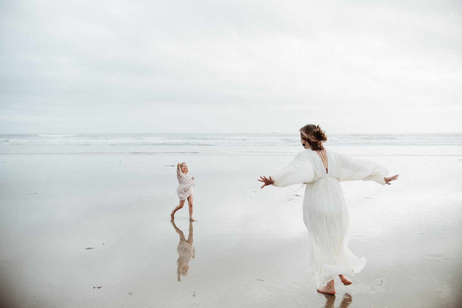 A woman and a young girl playing on a sandy beach with calm waves, both dressed in light-colored clothing, enjoying a cloudy day.