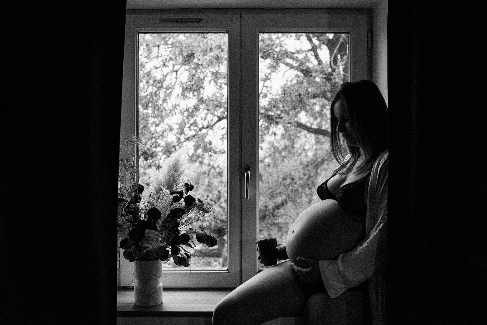 A pregnant woman sitting on the windowsill in her bedroom, holding a cup of tea and stroking her bump with her hand; her white blouse is open, and trees can be seen through the window; black-and-white photograph