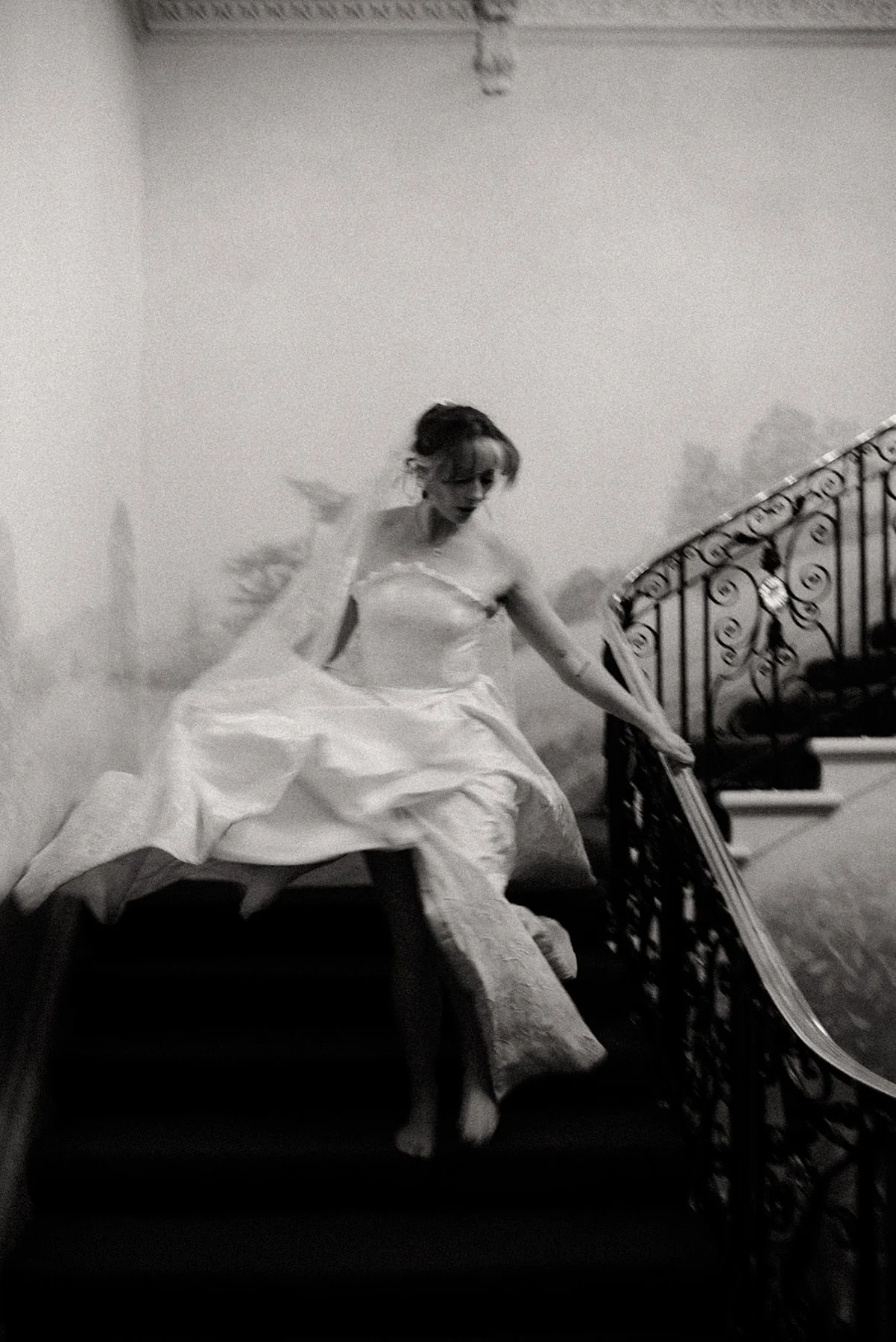 A bride running down the stairs, her dress billowing as she moves, revealing her bare legs and feet; a black-and-white, artistically blurred photograph taken in an English manor house in the Cotswolds