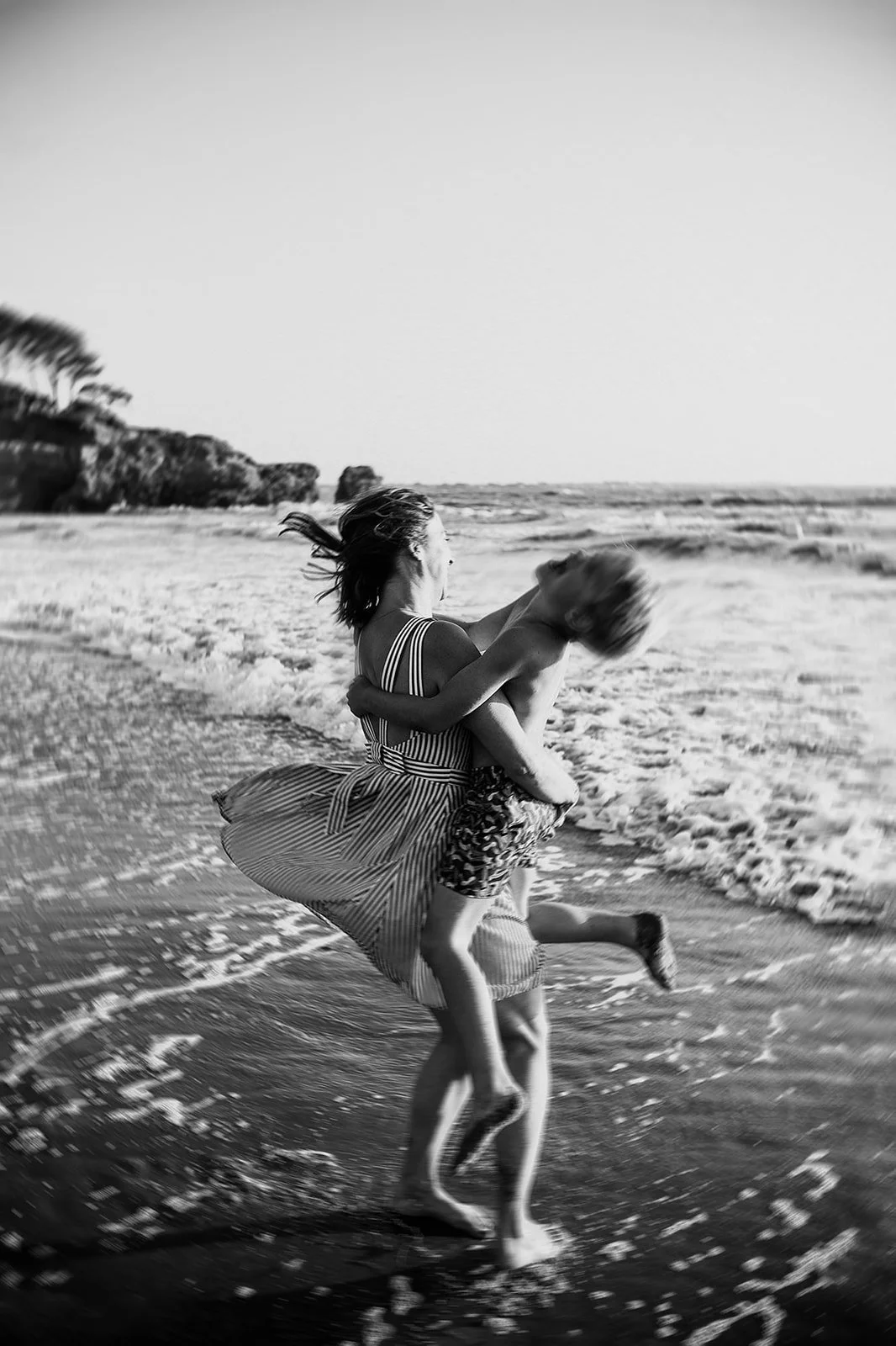 A mother and her son, who is jumping into her arms by the sea at the beach, with the waves crashing at their feet; her striped dress and brown hair blowing in the wind; a black-and-white photo with an artistic blur