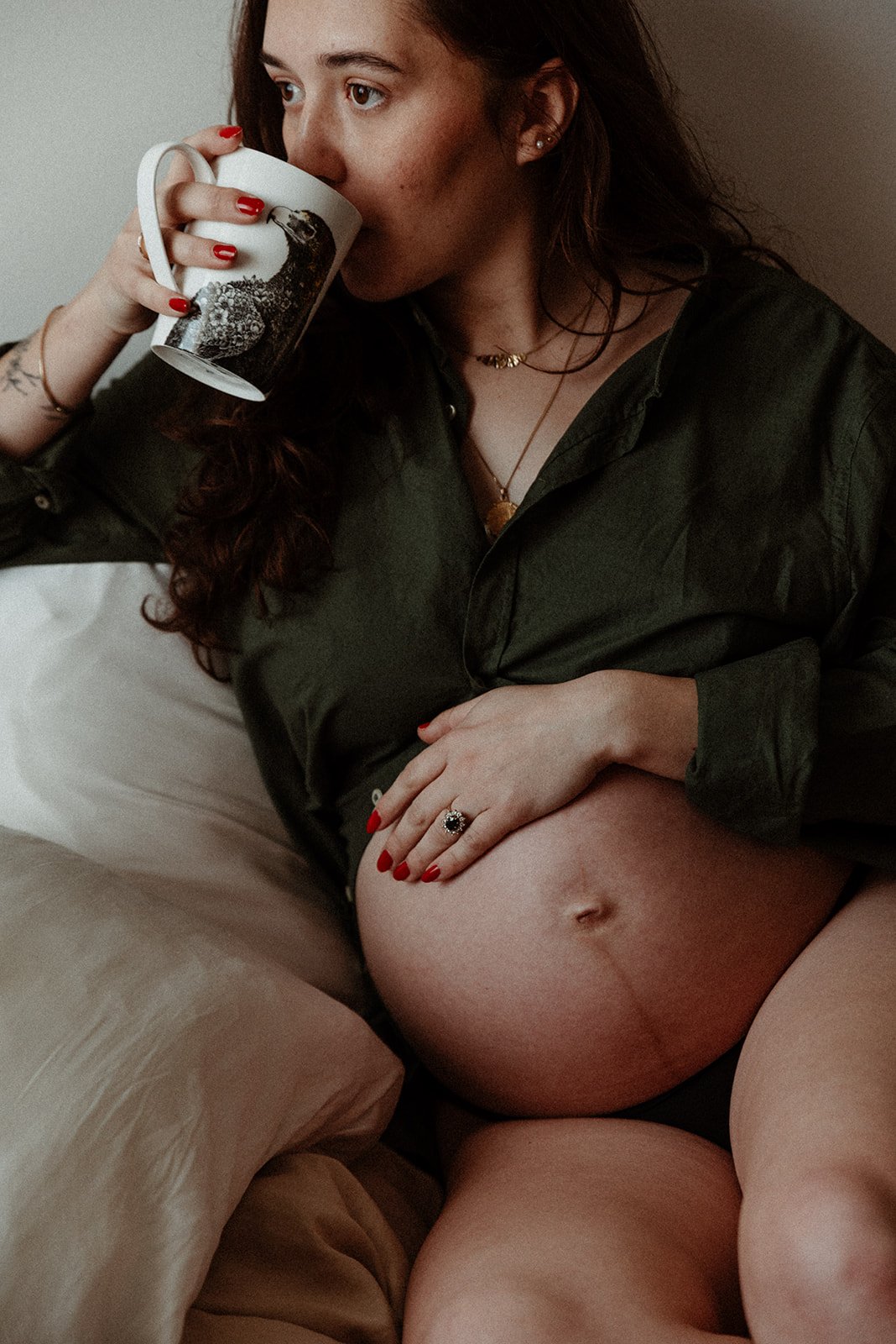 Portrait of a pregnant woman with long brown hair, wearing rings and necklaces, gazing right, drinking coffee in a mug, with her hand on her belly