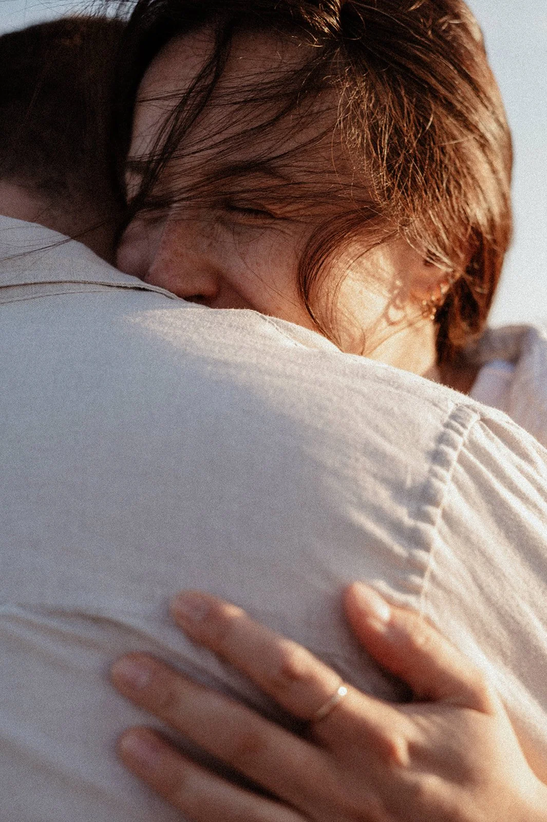 a close-up of a woman’s face, her eyes closed, her hair blowing in the wind, resting on her partner’s neck as they embrace