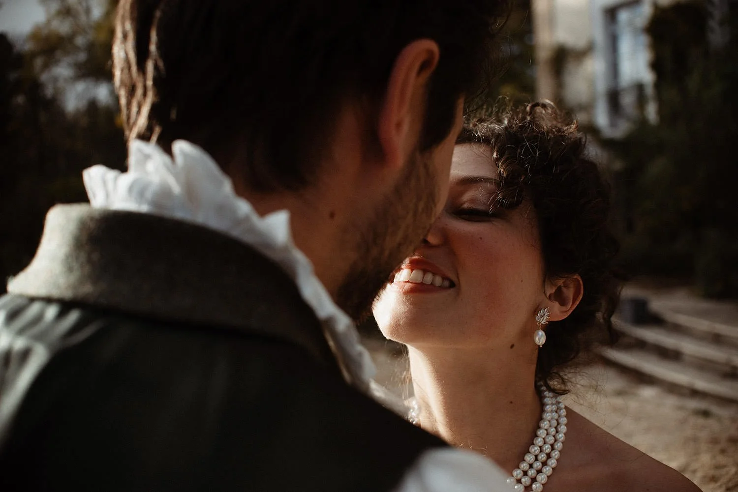 A couple sharing a close, intimate moment, with their faces near each other. The woman is smiling with closed eyes, wearing pearl jewelry and earrings. The man is wearing historical attire with a ruffled shirt and a coat. The background appears to be outdoors with stone steps.