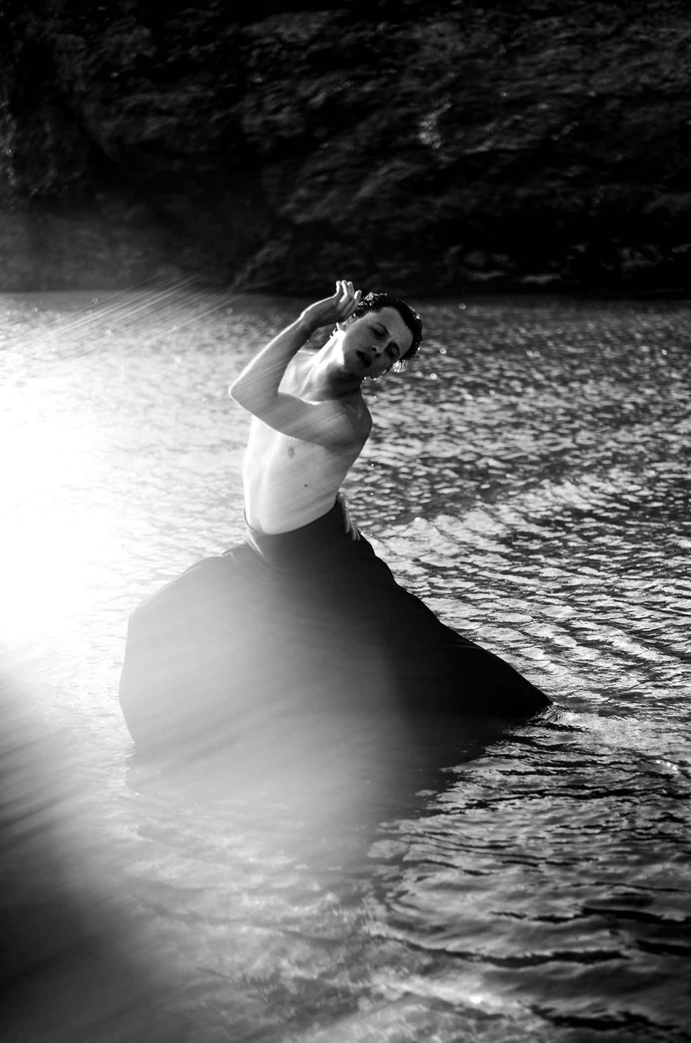 A black and white photo of a shirtless man sitting on a large rock in the water, with a rocky cliff in the background, raising one hand near his head.