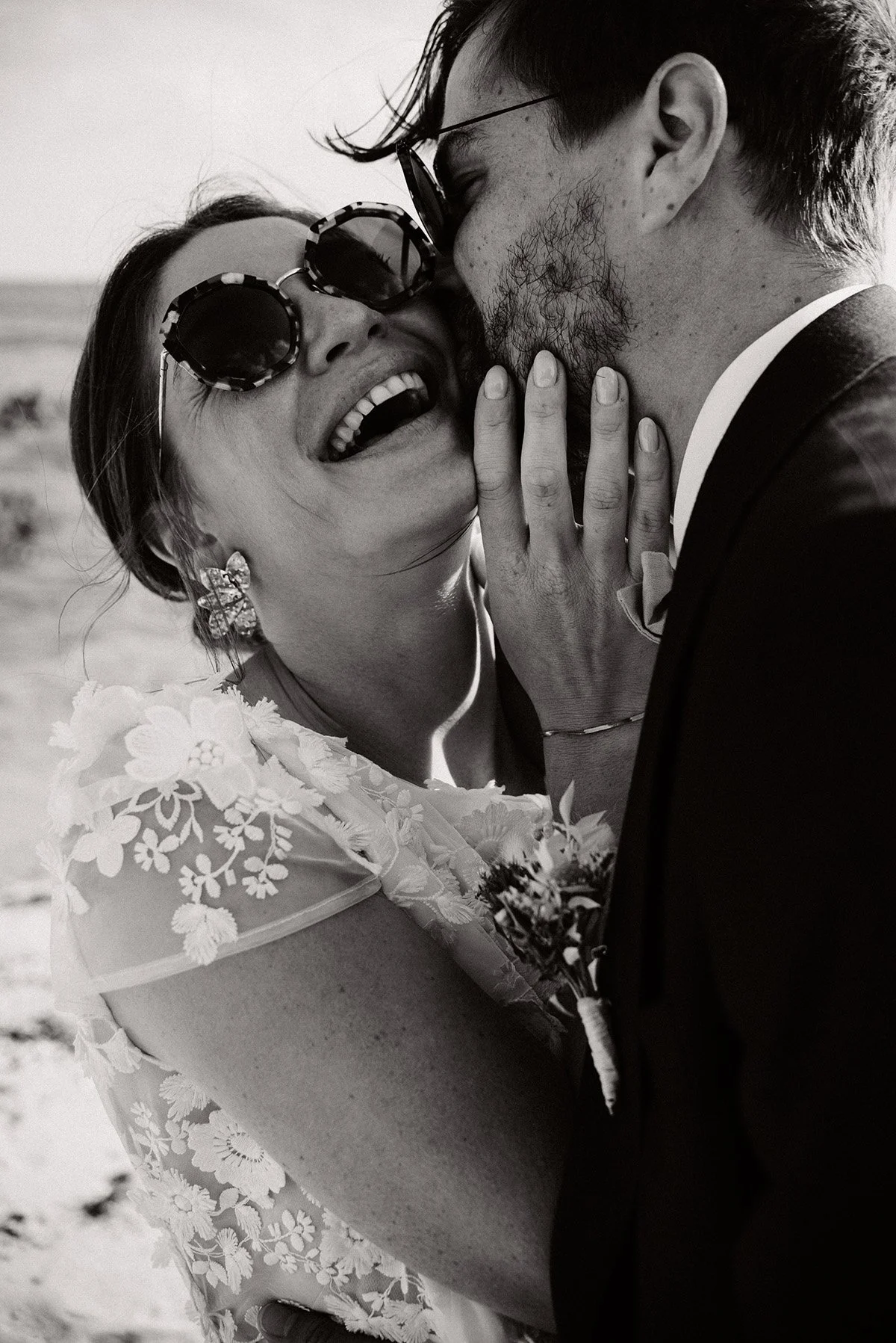 A black and white photo of a couple at their wedding. The woman is laughing and wearing sunglasses, a lace wedding dress, and flower earrings. The man, in a suit, is gently kissing her on the cheek while she touches his face.