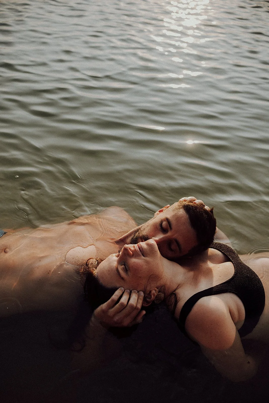 a photo of a couple in the water, floating on their backs, their heads resting on each other’s shoulders; he is kissing her on the cheek whilst holding her head
