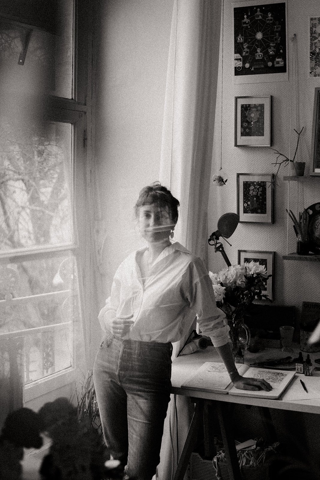 A woman with short hair, wearing a white shirt and jeans, standing near a window in her art studio decorated with framed artwork, a desk with books and a bouquet of flowers