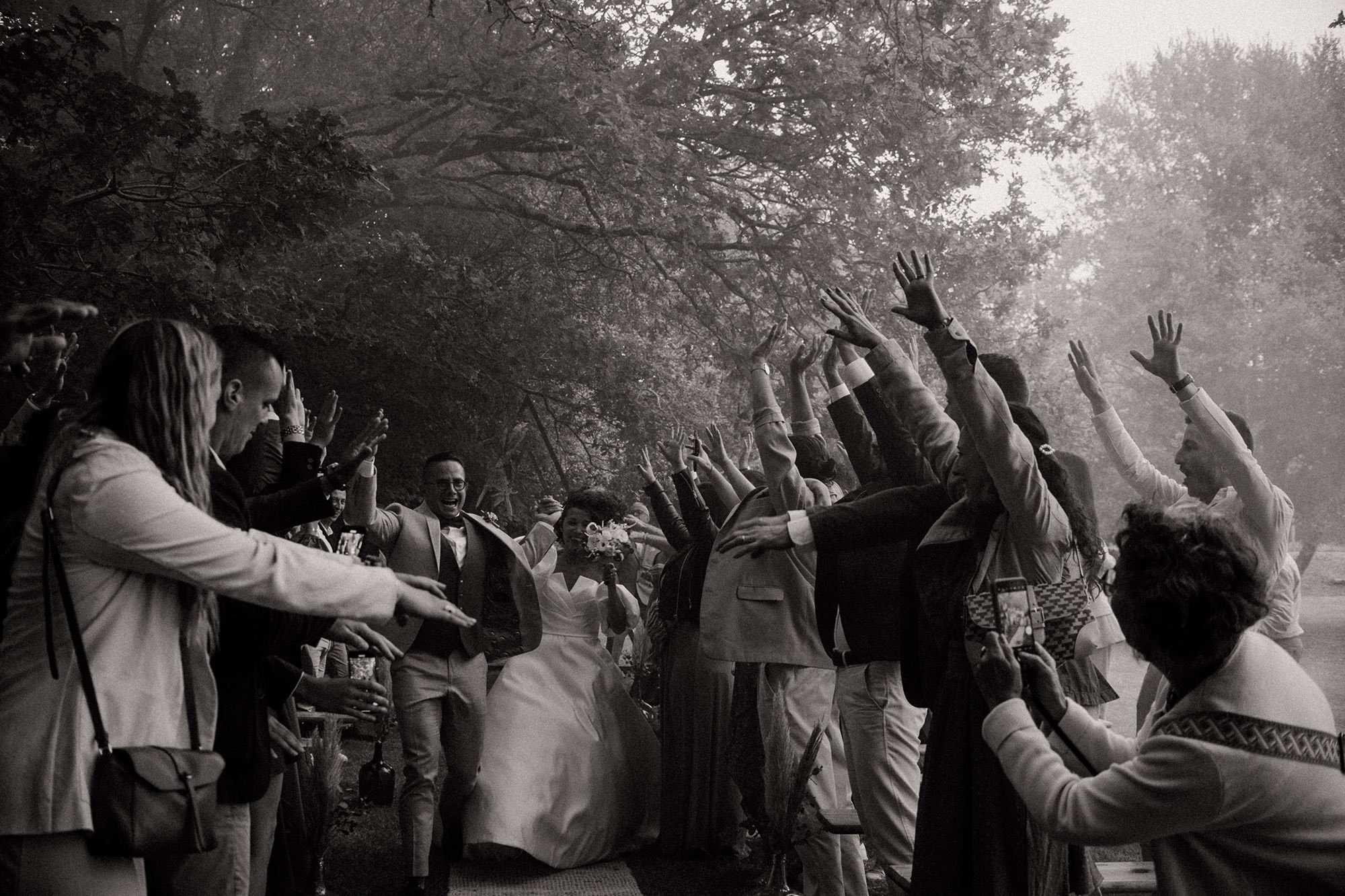 At the end of the secular ceremony under the trees, the bride and groom hold hands and run along the guard of honour formed by their guests, who have their arms raised, shouting cheers and setting off coloured smoke bombs