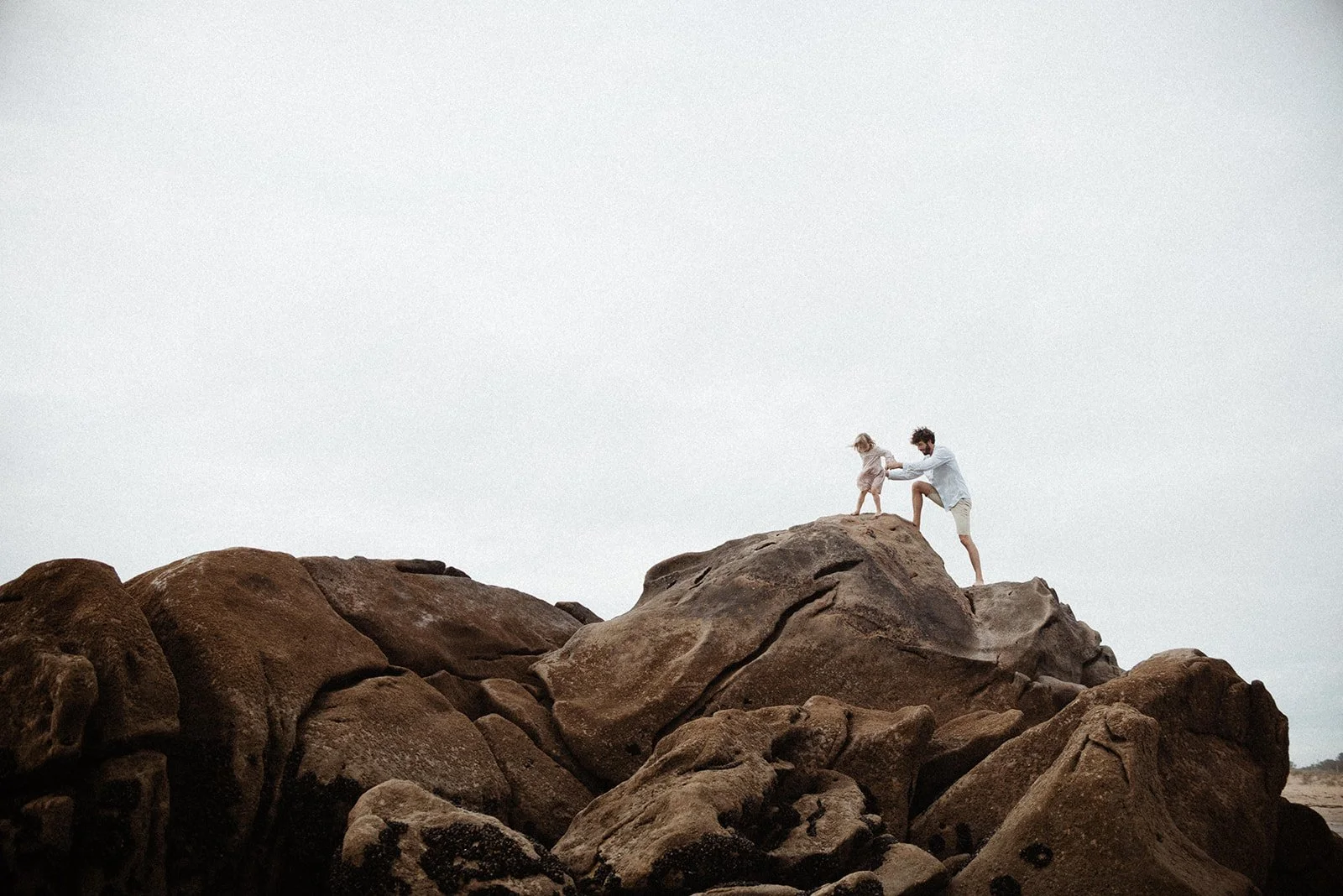 A father and his kid are climbing on large rocks at the beach, holding hands against a cloudy sky.