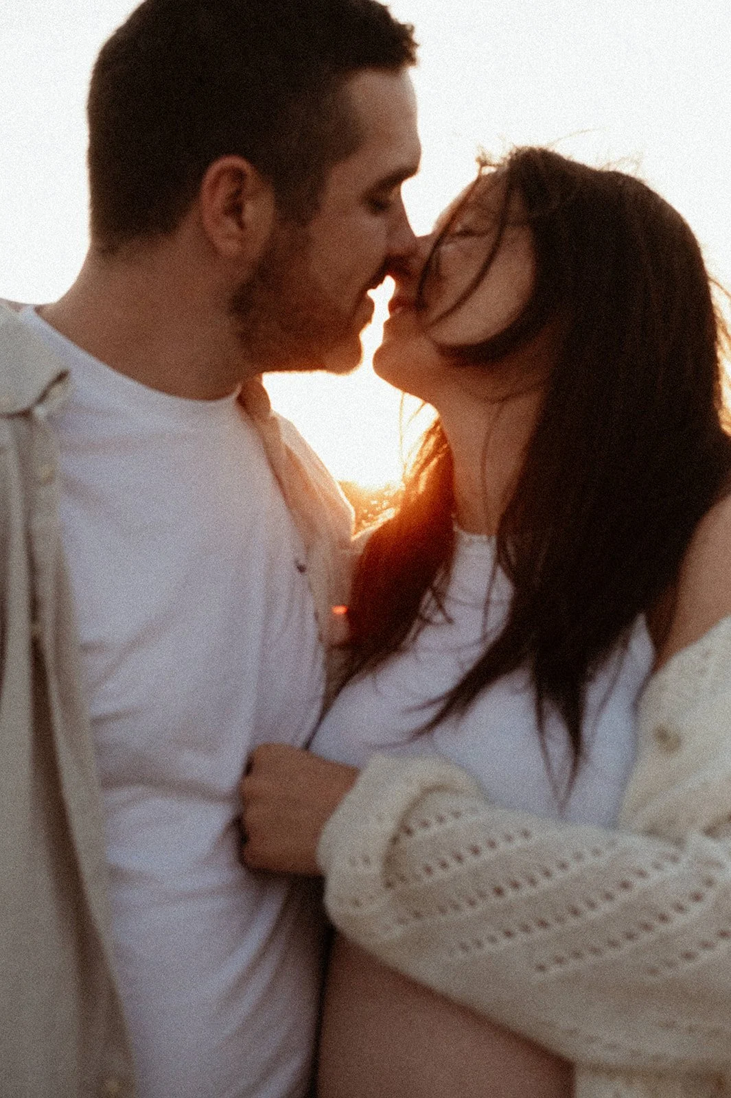 A close-up of a man and a long-haired brunette, their hair blowing in the wind, standing shoulder to shoulder, slowly leaning in to kiss, backlit by the setting sun between their faces, at golden hour